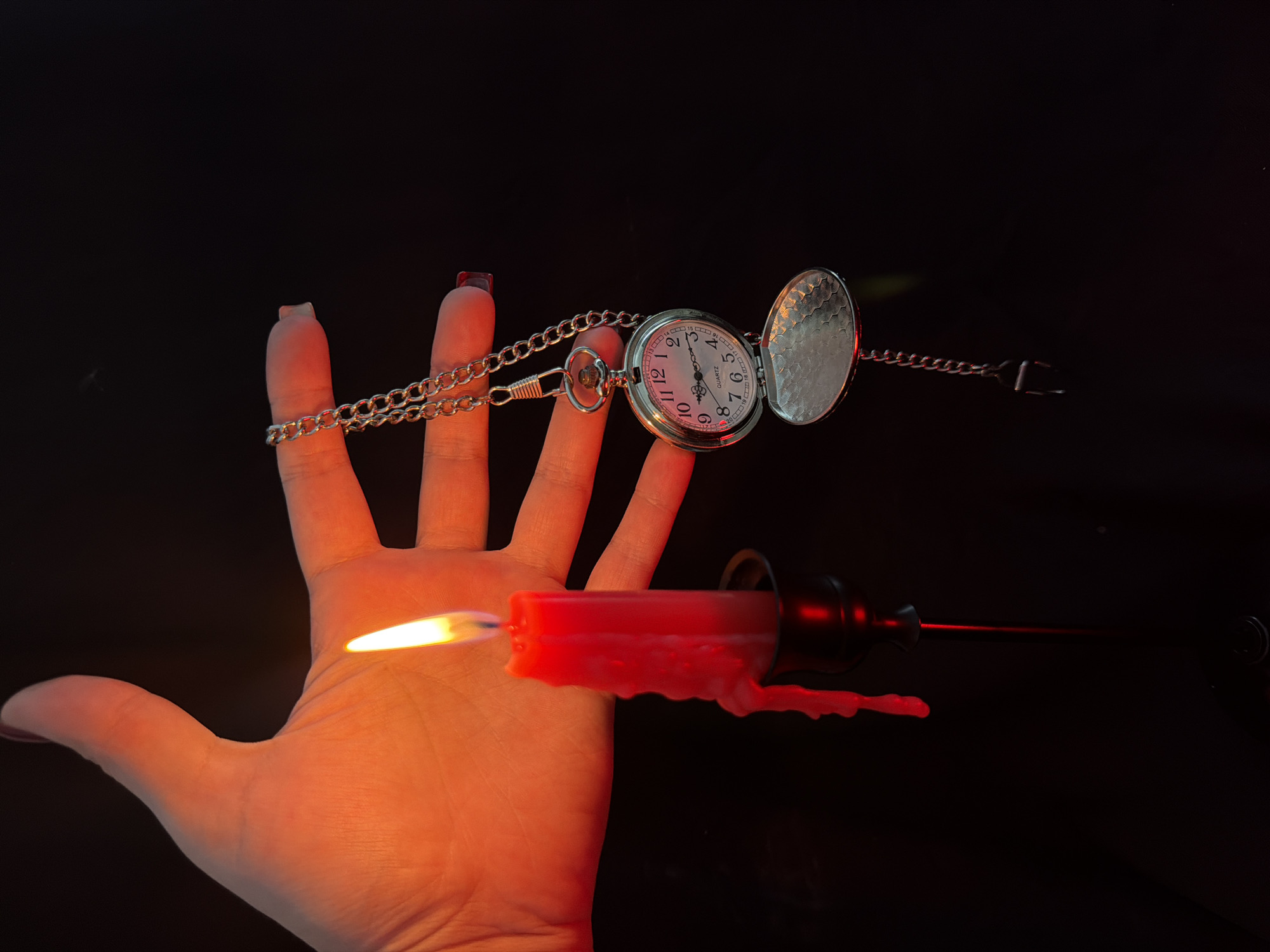A hand with red nail polish is held under a lit red candle, its flame casting an orange glow on the palm. A silver pocket watch on a chain hangs from the hand, its face open to reveal a white clock face with black Roman numerals. The watch is illuminated by the candle's light. A black candlestick holder is visible below the candle. The background is completely black.