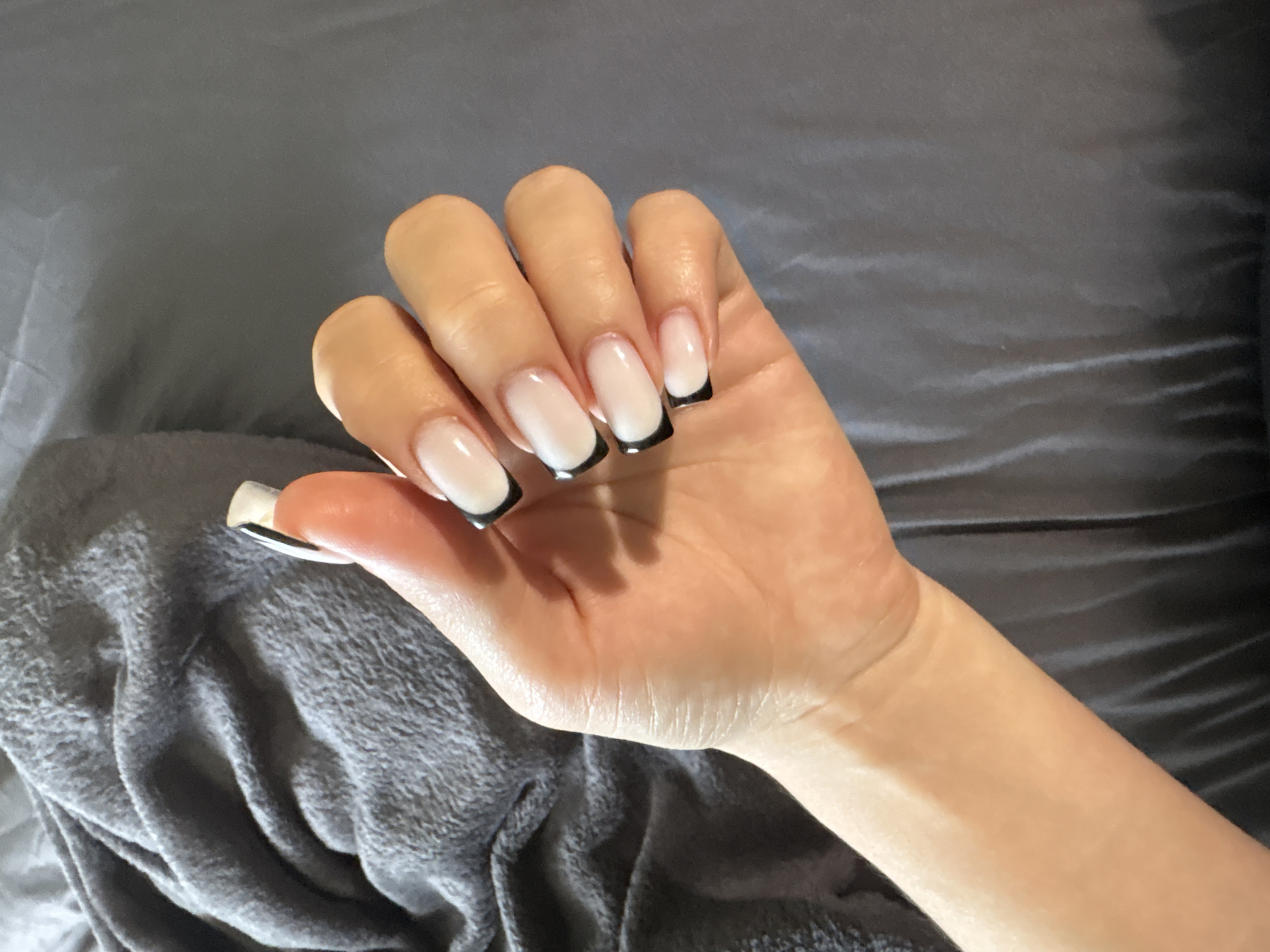 A close-up overhead shot shows a person's left hand with its fingers curled against a dark grey, wrinkled fabric. The hand has several nails that are painted with a French manicure style, featuring a sheer pale pink base and a sharp black tip. The fabric appears to be a blanket or throw. There is no identifiable location in the city of Rezh, Russia.