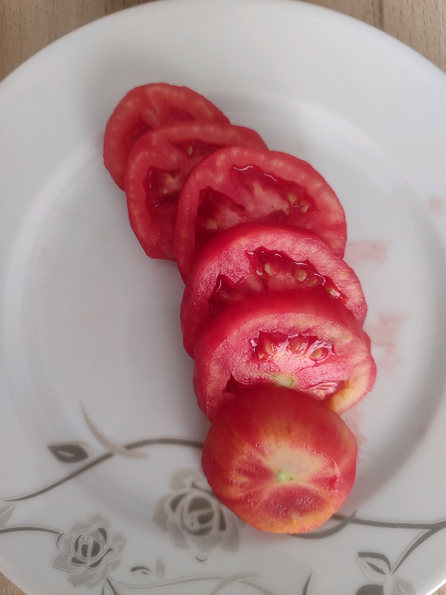A close-up, high-angle shot shows five sliced tomatoes arranged diagonally on a white plate with a subtle floral pattern. The tomatoes are a vibrant red, with visible seeds and juice glistening on their surfaces. The bottom-most slice is whole, while the others are cut into thin rounds. The plate rests on a textured surface, possibly a wooden table, which is visible in the upper portion of the image. The lighting is natural and even, casting soft shadows. The scene is static and appears to be a still life composition. No people or specific location details in Gümüşova, Turkey are discernible.