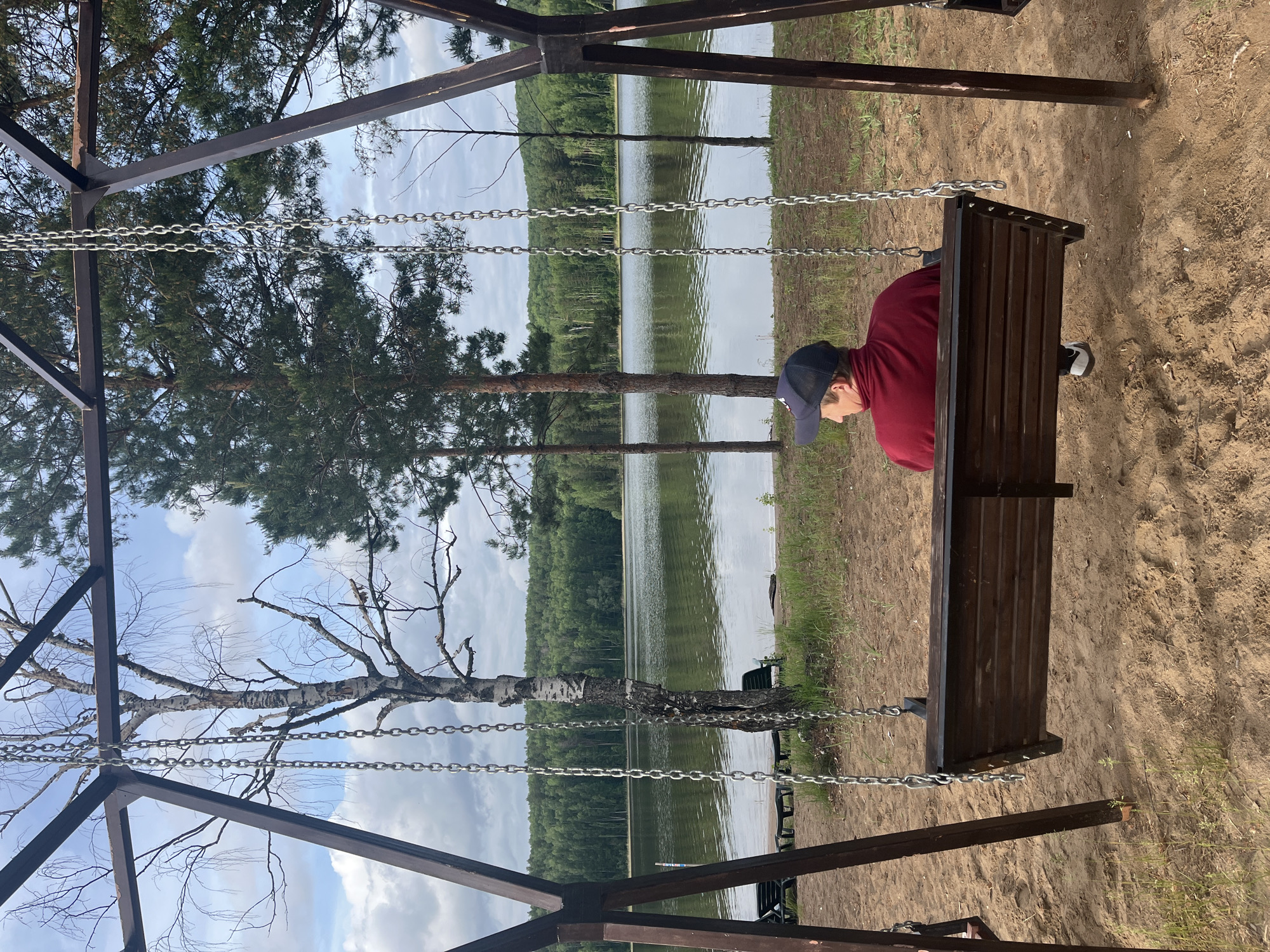 A person in a maroon shirt and a blue baseball cap sits on a wooden swing-bench facing away from the viewer. The bench is suspended by chains attached to a dark-colored metal frame structure. The ground surrounding the swing is sandy. Beyond the swing, a lake is visible with trees lining the opposite shore. The sky is partially cloudy. The location is Unknown, Unknown.