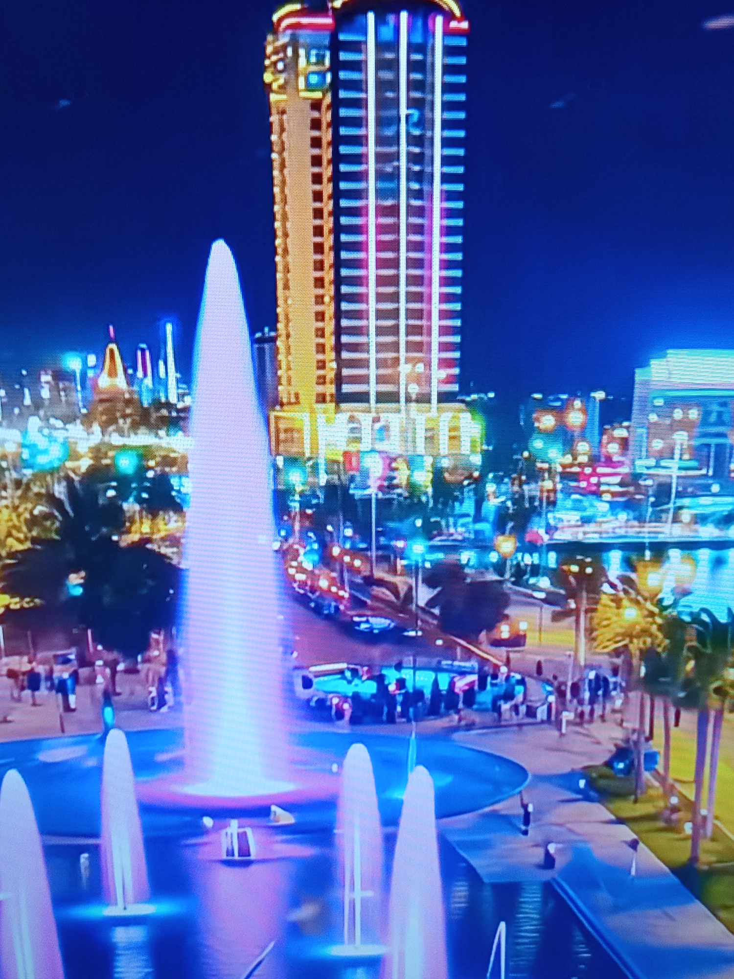 The image depicts a nighttime view of a city skyline with a prominent skyscraper illuminated by a grid of colorful lights. In the foreground, a large, illuminated fountain sprays water upwards, casting a pink and blue glow. Several smaller fountains are also visible. The background shows a cityscape with numerous buildings, streetlights, and some vegetation, including palm trees. People are visible walking along a pathway and gathered near vehicles in the middle ground. The scene appears to be a public plaza or waterfront area within a city. The location is not Brno, Czechia.