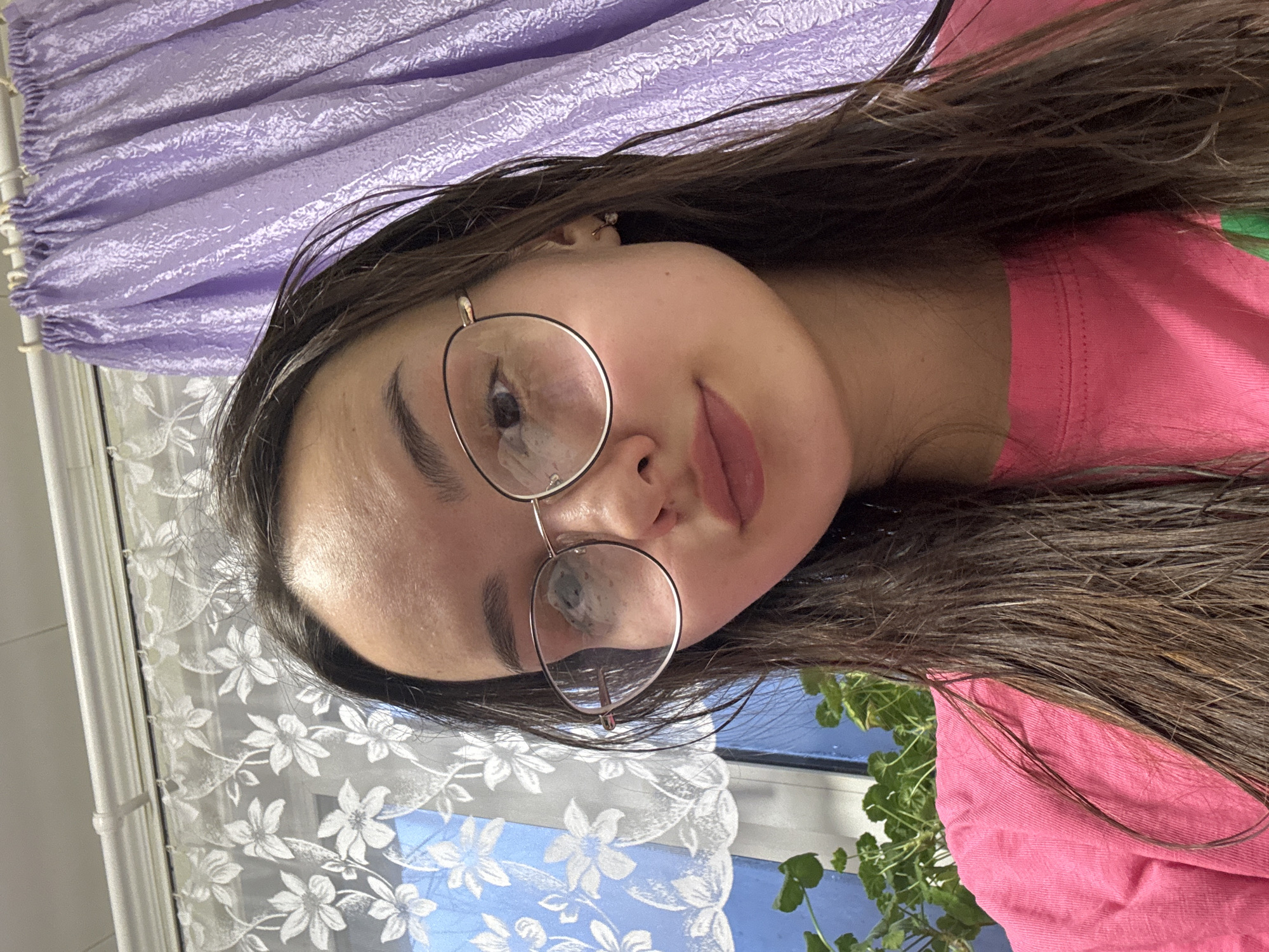 A young woman with brown hair and wearing round glasses is the subject of the image. She is wearing a pink shirt. The background includes a purple curtain, a window with a white floral lace curtain, and a glimpse of a plant outside the window. The location in the city of Urada, Russia, cannot be determined from the image.