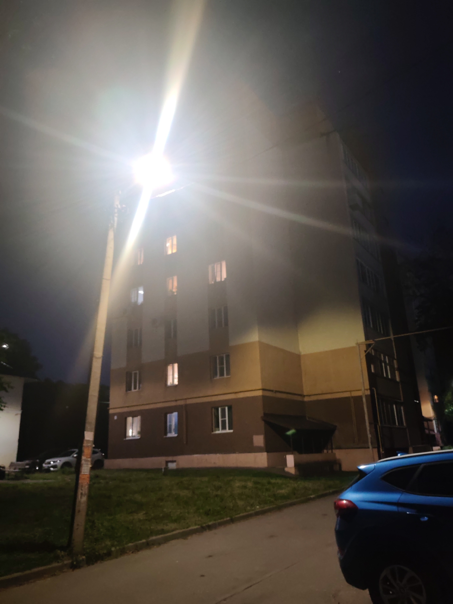 The image captures a nighttime street scene in the city of Vladimir, Russia, near a multi-story residential building. A bright streetlamp, positioned to the left of the frame, illuminates the foreground and casts strong light flares. The building, with its cream-colored upper facade and brown lower section, has several illuminated windows, suggesting occupancy. A blue SUV is parked on the right side of the frame, facing away from the camera. Adjacent to the building is a grassy embankment. The overall impression is of a residential area at night, with artificial lighting creating a dramatic contrast with the dark sky.