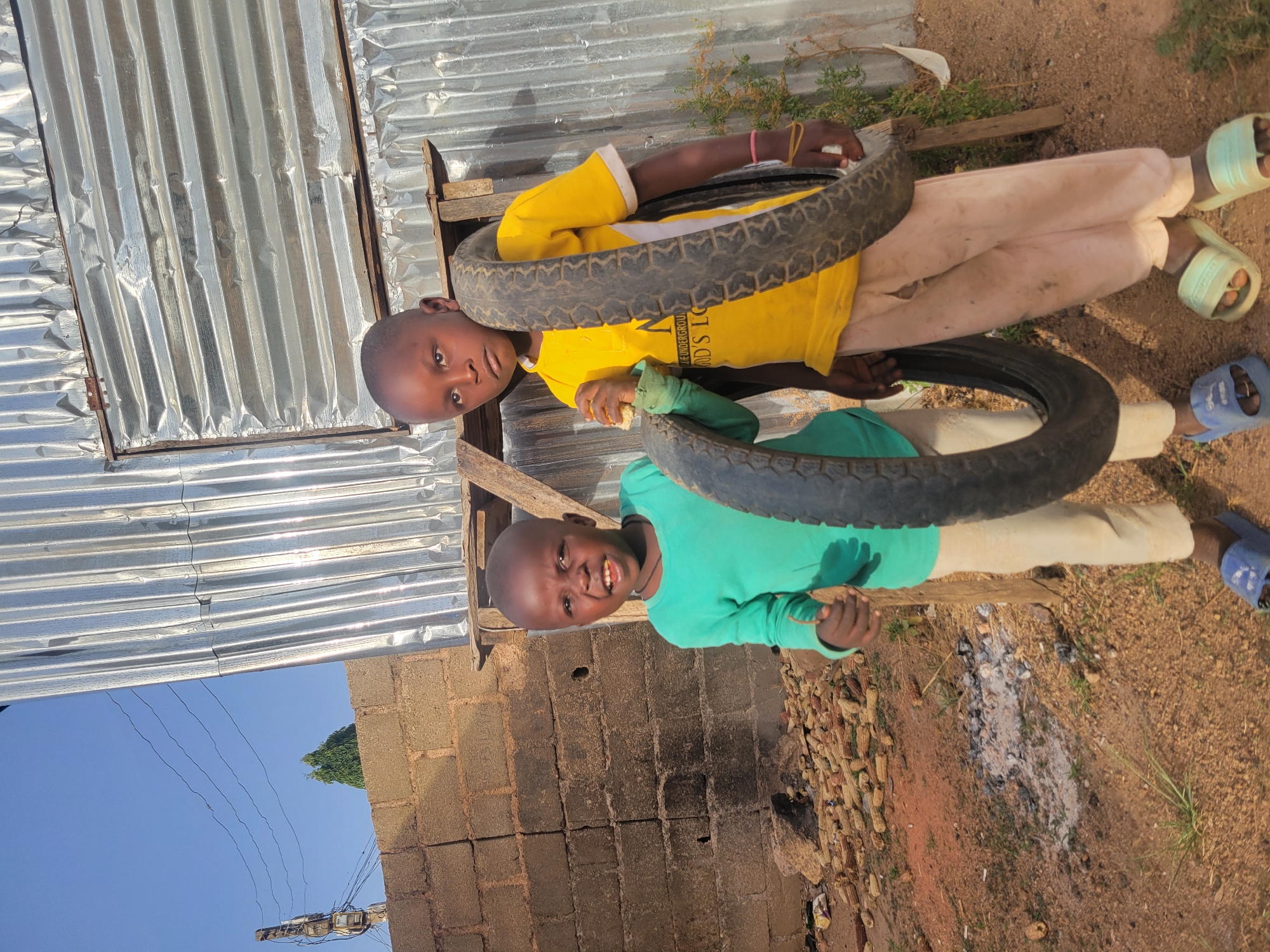 This image, taken in Barakin Lerawi, Nigeria, depicts two young boys outdoors in bright daylight. The scene is captured at a 90-degree counter-clockwise rotation from standard viewing.
The boy on the left (or top, in the rotated image) has a shaved head and wears a bright yellow long-sleeved t-shirt, light-colored pants, and light green slide sandals. A dark tire encircles his midsection. He looks directly at the camera with a neutral expression. Visible text on his yellow shirt includes fragmented words like "...THE UNDERGROU..." and "...OD'S STO...".
Beside him, the second boy, also with a shaved head, wears a teal long-sleeved t-shirt, light-colored pants, and blue slide sandals. He is smiling broadly at the camera, revealing his teeth, and is also holding or playing with a dark tire.
The background features a structure made of corrugated metal sheets, which are shiny and reflect sunlight. Below this, a wall constructed from dark concrete blocks is visible. The ground is reddish-brown dirt with some sparse green vegetation. A pile of dried corn cobs lies on the ground near the smiling boy. The sky is clear blue, with visible power lines, a utility pole, and a distant green tree, indicating an outdoor, possibly rural or semi-urban setting. The children appear to be playing or posing for the camera with the tires.
