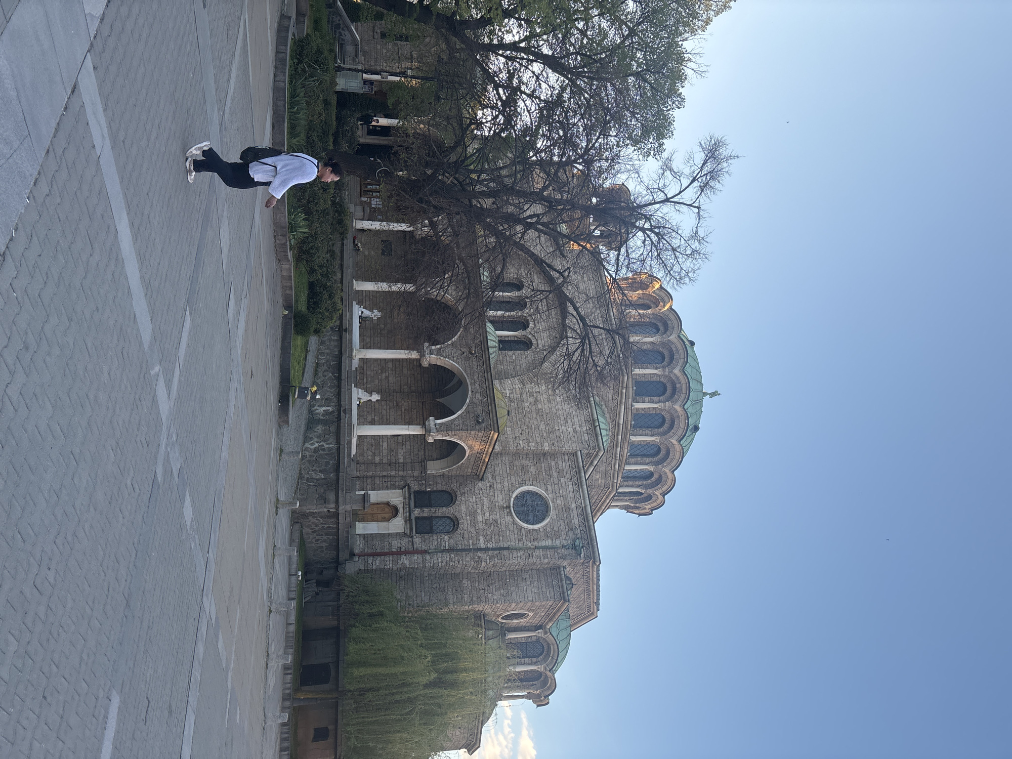 A medium shot shows the St. Alexander Nevsky Cathedral in Sofia, Bulgaria. The church is made of light-colored stone and has a large dome and several smaller domes. A bare tree is situated in front of the church, its branches partially obscuring the view of the building. A person dressed in white shirt, black pants and white shoes is walking on a light-colored stone surface. The sky is clear and blue.
