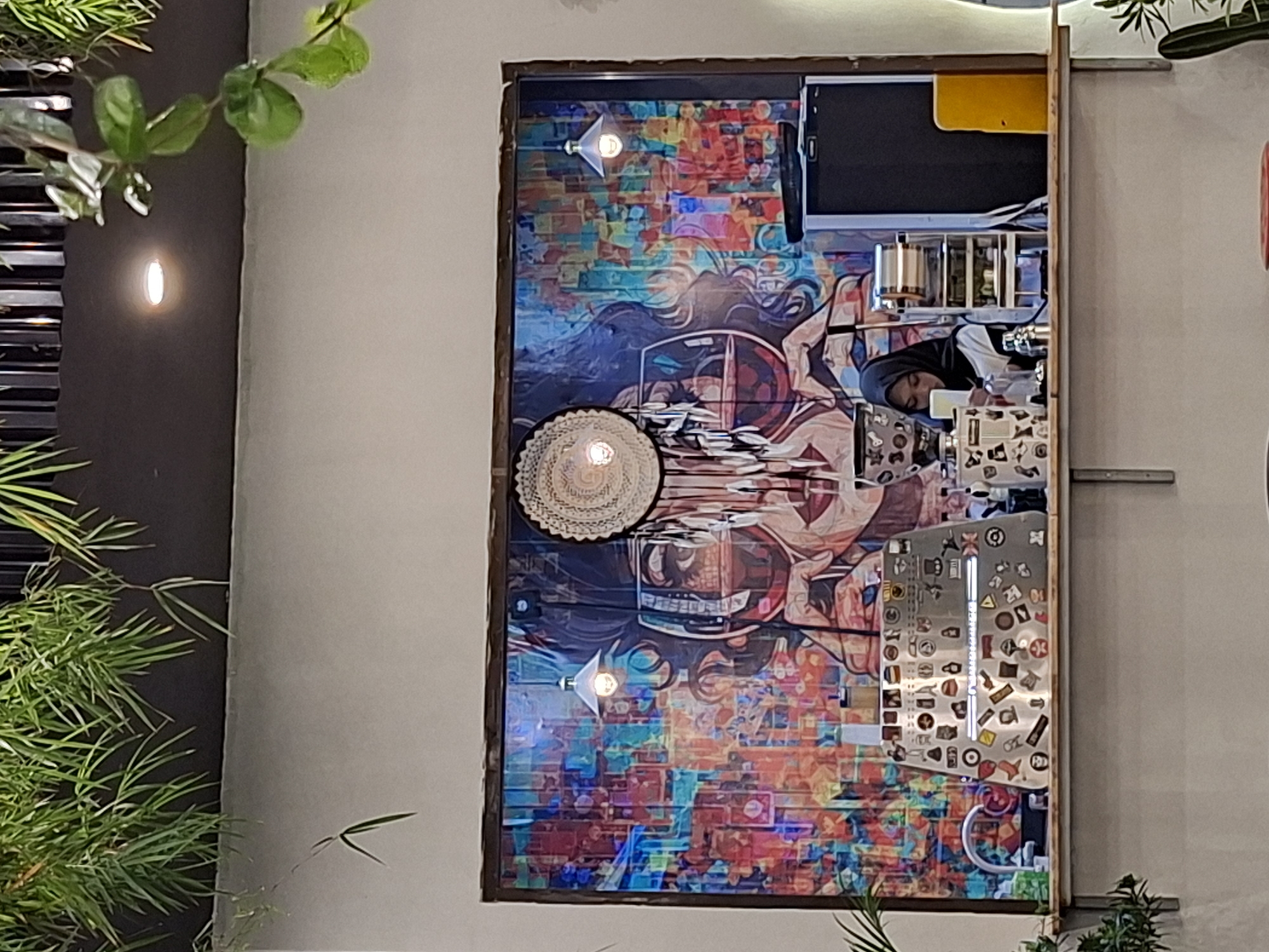 Here is a detailed description of the image:
**Scene Overview:** The image captures a frame of an establishment, possibly a cafe or restaurant, in what appears to be Lhokseumawe, Indonesia, based on the context.
**Subjects and Objects:**
- A large, framed piece of art dominates the center. The artwork is colorful and appears to be a modern, abstract piece with incorporated imagery of people and urban elements.
- A woven, flat disc with a lightbulb inside sits at the center of the artwork.
- Reflecting the artwork is the reflection of a person in a headscarf working in the reflected cafe or restaurant.
- Several objects related to a food preparation area are visible, including metal containers and what appears to be equipment for food or beverage preparation.
- The entire setup is framed by a building's exterior, with plants (likely bamboo or similar foliage) visible on the sides and in the background, adding a natural element to the urban setting.
**Environment:** The environment is a blend of indoor and outdoor, with the establishment seemingly having an open or partially open structure. The lighting suggests it's either daytime or the interior is well-lit. The presence of plants indicates an effort to incorporate nature into the urban setting.