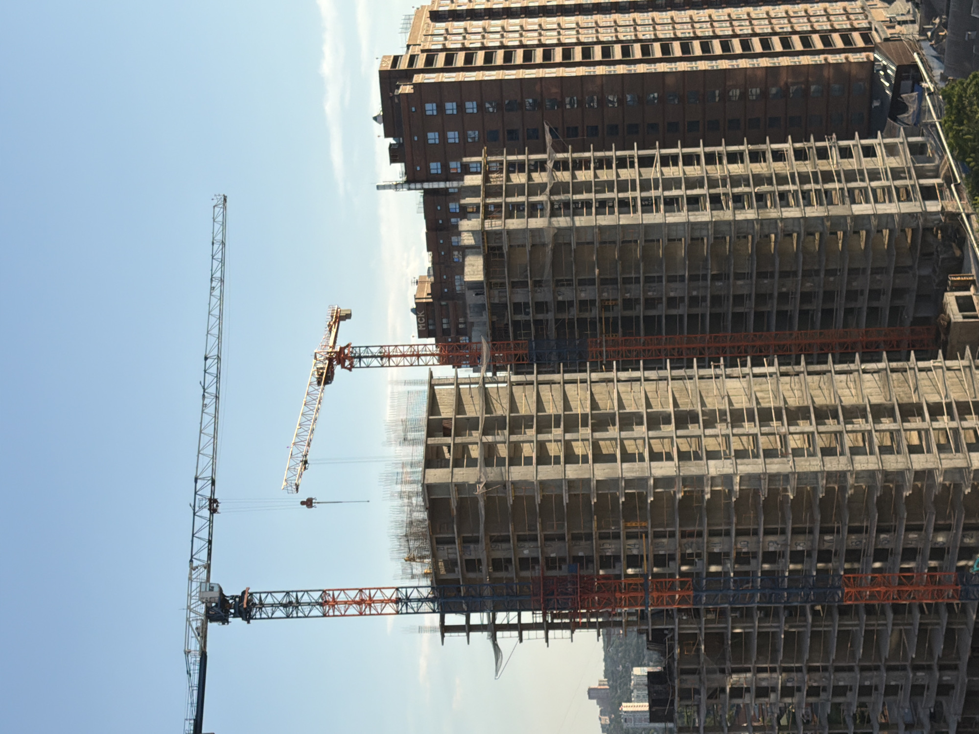 In Ovoshchnoy, Russia, two towering construction cranes are positioned against a clear blue sky, one on the left with a red and blue lattice structure and a long horizontal boom, and another to the right with a red boom extending outwards. In the foreground, unfinished multi-story buildings with exposed concrete frameworks are under construction, featuring multiple levels of visible floor slabs and rebar. A completed, taller, reddish-brown building with many windows stands to the right of the construction site. Below the main construction area, various construction materials and debris are visible, along with some trees and a small structure that appears to be a temporary shelter. The scene depicts an active urban development project.