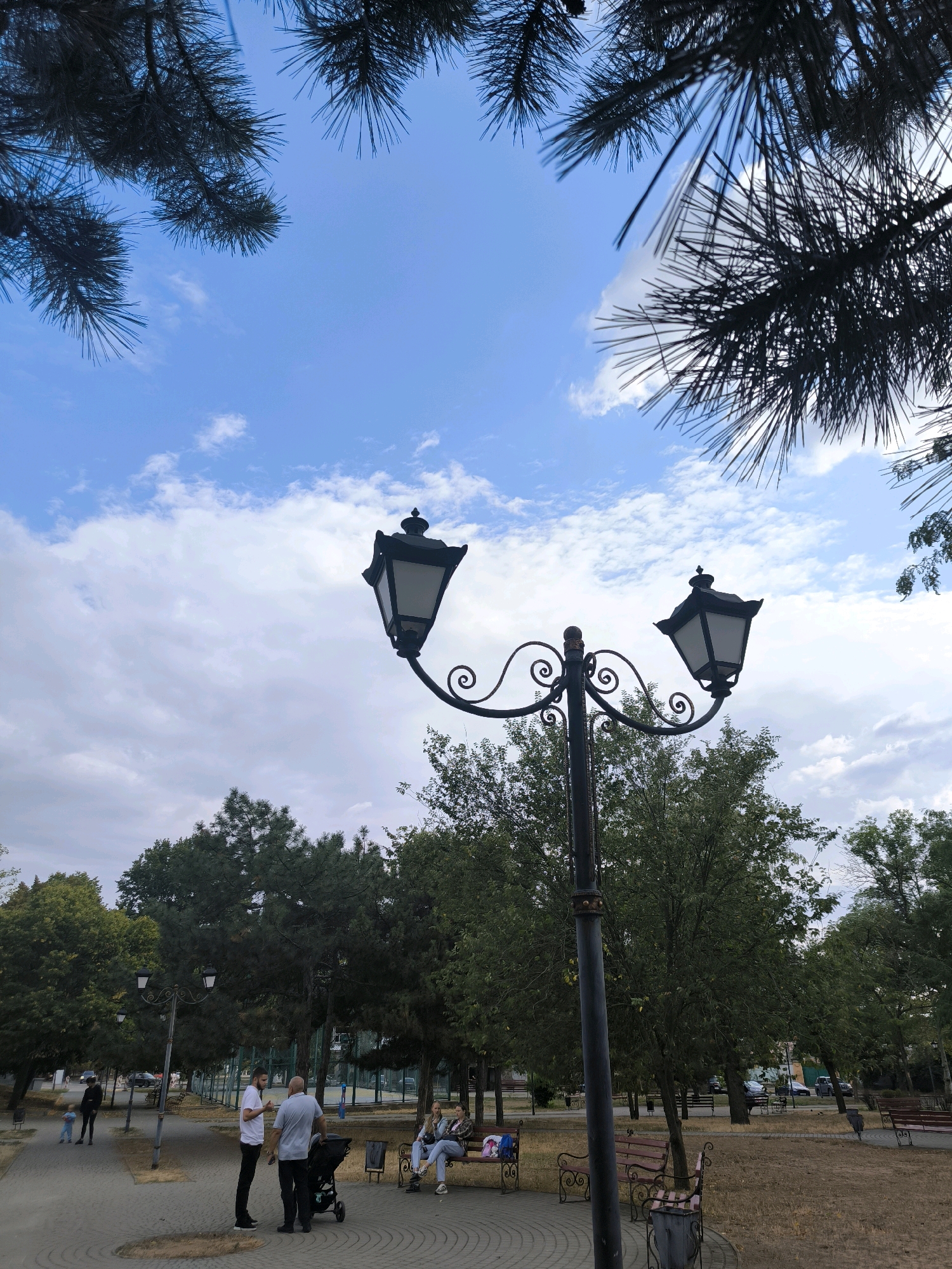 The image captures a view of a park in Mykolaiv, Ukraine, under a cloudy blue sky. In the foreground, a decorative black lamppost with two lanterns stands prominently. Pine branches frame the top of the image, silhouetted against the sky. In the middle ground, several people are present. Three individuals stand and converse near a baby stroller on a paved walkway. Further to the right, two women are seated on a park bench. Behind them, more park benches and trees are visible. In the background, a sports field with a fence is discernible, and beyond that, buildings and parked cars. The overall environment appears to be a public urban park during daylight hours.