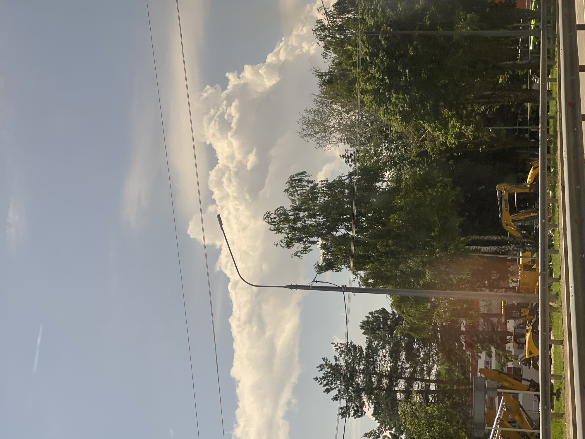 The image shows a daytime scene in Верхнерусское, Russia, under a partly cloudy sky. The sky is a bright blue with large, fluffy white clouds dominating the left side of the frame. The sun is shining, casting a warm light on the scene.
In the foreground on the right, a metal guardrail with horizontal yellow and gray bars suggests a roadside or construction area. Beyond the guardrail, a line of lush green trees, including deciduous and coniferous varieties, forms a dense backdrop. The trees are vibrant and full, indicating a warm season.
Several yellow construction vehicles, likely excavators or backhoes, are visible among the trees and in the middle ground. Some are parked, while others appear to be in operation, suggesting ongoing construction or land development. A tall lamppost with a curved arm stretches across the sky from the lower left towards the center, its shadow faintly visible. Power lines crisscross the sky, adding to the urban or semi-urban feel of the location. In the background, a building with a light-colored facade and red accents can be seen peeking through the trees, hinting at a residential or commercial area.
The overall impression is one of a bright, active day in a developing or undergoing maintenance area, with nature and construction coexisting. There is no visible text in the image.
