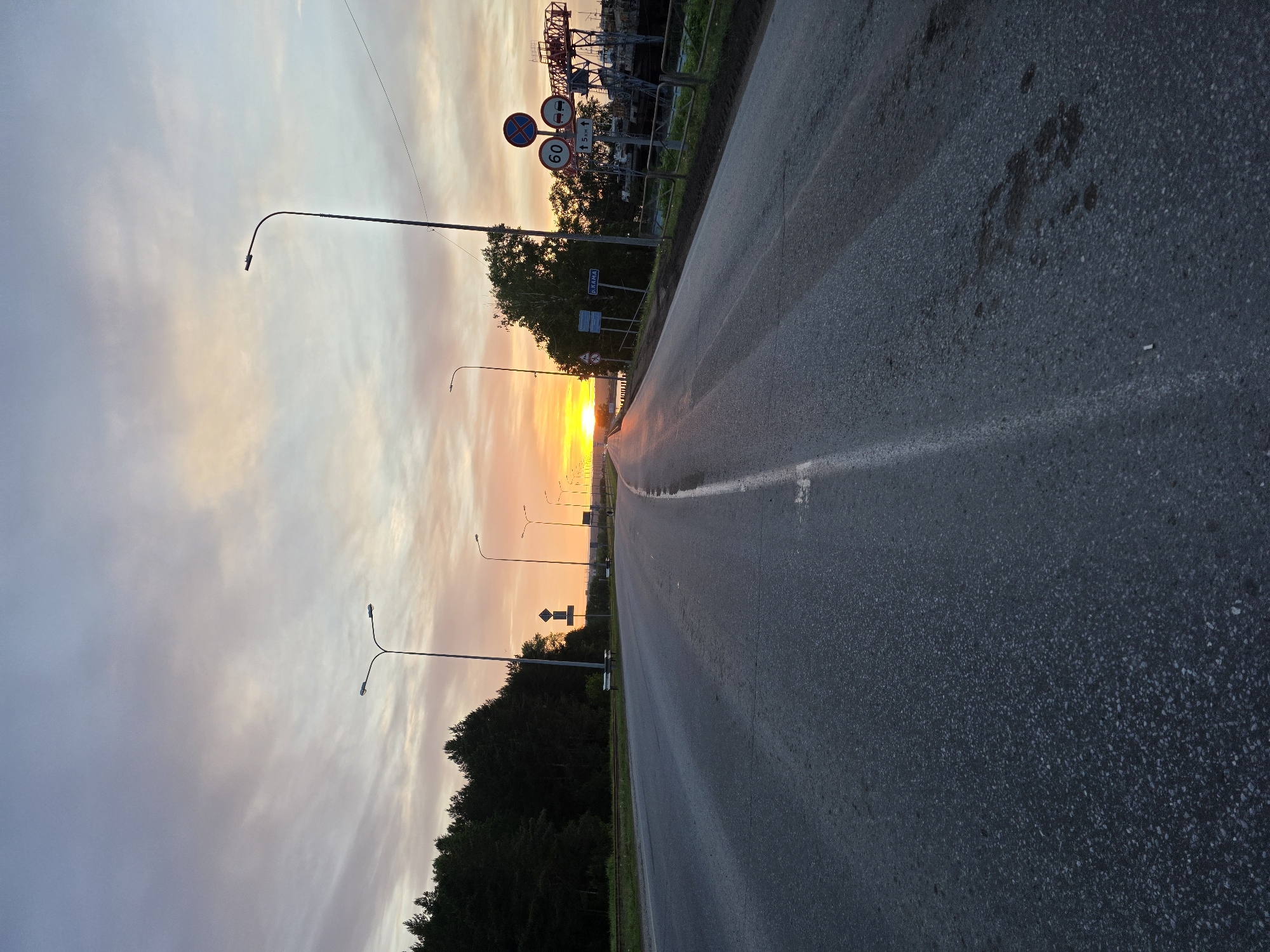 This image captures a scene on a road in Chaikovsky, Russia, during what appears to be a sunset. The road is asphalt, showing signs of wear. A single white line marks the road's edge. Streetlights line the left side of the road, spaced at intervals. Beyond the streetlights, trees are visible. The sky is filled with clouds and the sun is setting on the horizon. Road signs are located on the right side of the road. In the background, structures are visible. The perspective is at street level. There are no people in the image.