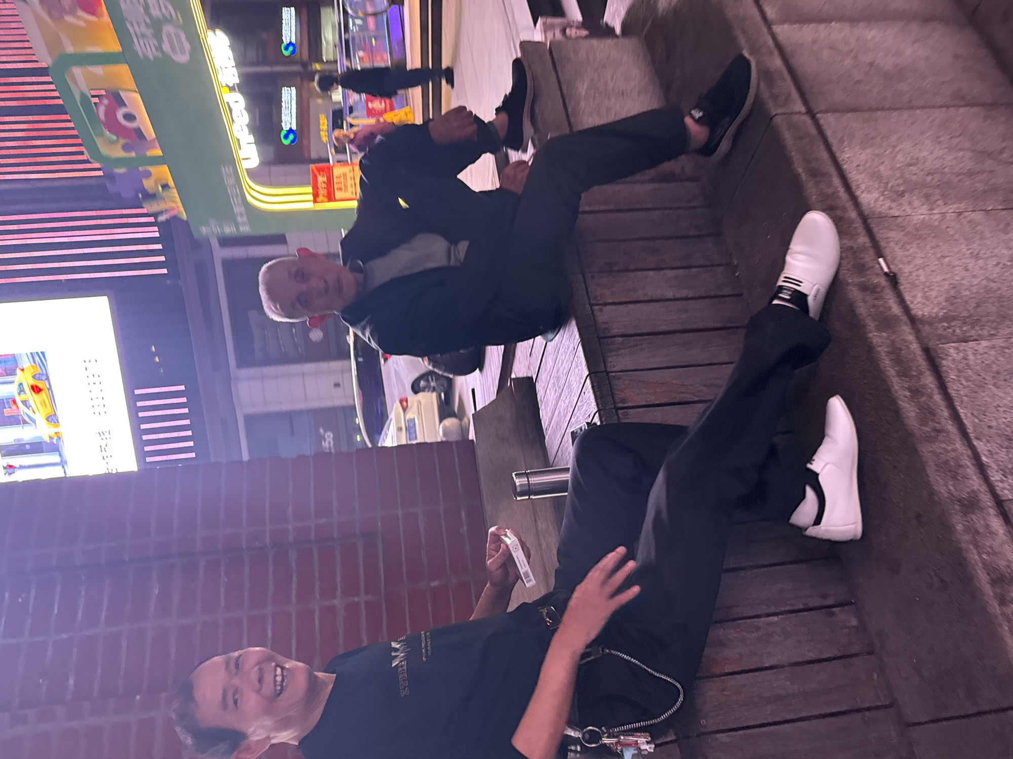 Two men are sitting on a wooden bench outdoors in Yuzhong District, China. The man on the right is older, wearing a black t-shirt and pants, holding a pack of cigarettes. The man on the left is wearing a gray shirt and black jacket and pants. Bright, colorful lights illuminate the urban background.