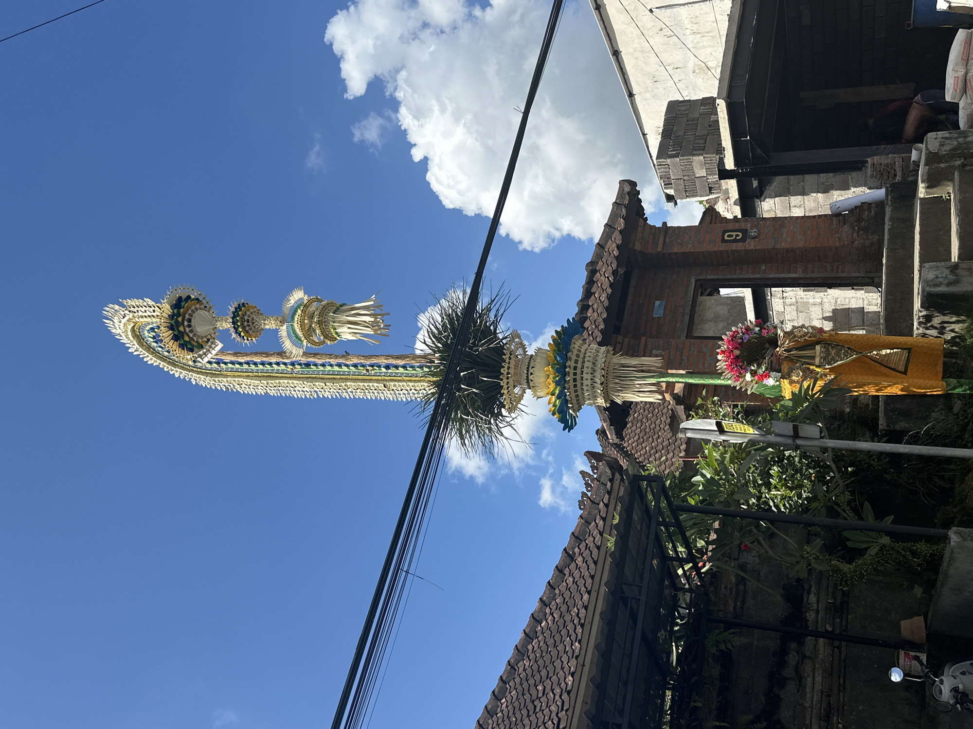This is a medium shot of a street scene in Ubud District, Indonesia, on a bright, sunny day with a blue sky and scattered clouds. The focal point is a tall, ornate Balinese decoration, likely a Penjor, made of woven materials, palm fronds, and colorful accents of yellow, blue, and green. It stands upright against the sky, partially obscured by overhead power lines.
In the foreground, to the left, is a section of a traditional tiled roof of a building with a dark overhang. Further left, a motorcycle is parked, and a wall covered in creeping plants is visible.
To the right of the Penjor, a brick gateway with a number "9" is prominent. Steps lead up to this gateway, which appears to be part of a building under construction or renovation, evidenced by stacks of grey bricks and exposed building materials. Shadows suggest the sun is high in the sky.
A person is partially visible in the lower right corner, near what appears to be bags of cement, indicating ongoing construction work. The overall impression is a blend of traditional Balinese culture and the ongoing activities of daily life and development in a tropical urban setting.