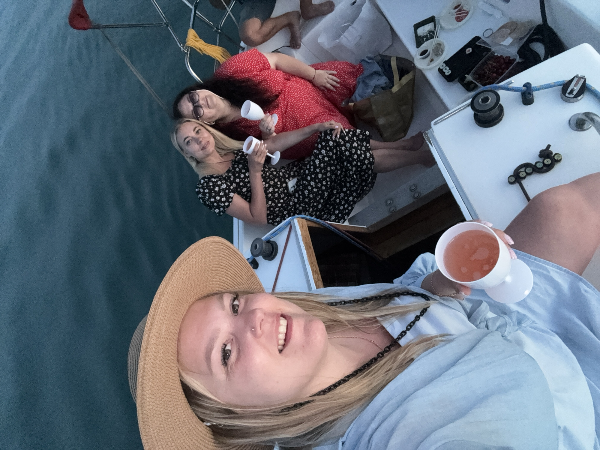 The image depicts three individuals on a boat during what appears to be dusk or evening, with a partial figure also visible. In the foreground, a light-skinned woman with blonde hair and a nose piercing wears a straw hat, a light blue long-sleeved top, and a dark chain necklace. She is smiling at the camera while holding a white goblet containing a pink beverage.
To the center-right, a second light-skinned woman with blonde hair wears a black dress with white floral patterns. She is also smiling at the camera and holding a white goblet with a clear or light-colored beverage. To her right, a third light-skinned woman with dark hair wears sunglasses and a red dress with white polka dots. She is also smiling at the camera, holding a white goblet with a clear or light-colored beverage. A fourth individual's bare legs and feet are partially visible on the far right side of the boat.
The boat's deck is white, furnished with marine hardware including winches, cleats, and metal railings. A blue line is coiled near a winch, and red and yellow fabrics are draped over a railing. A brown bag, small containers, and plates with various items, including what appear to be berries, are positioned on the deck behind the two women in the background. The surrounding water is dark blue-green with minimal surface agitation. The scene is illuminated by low light conditions, consistent with evening. This event is stated to be located in the city of Krasny, Russia.