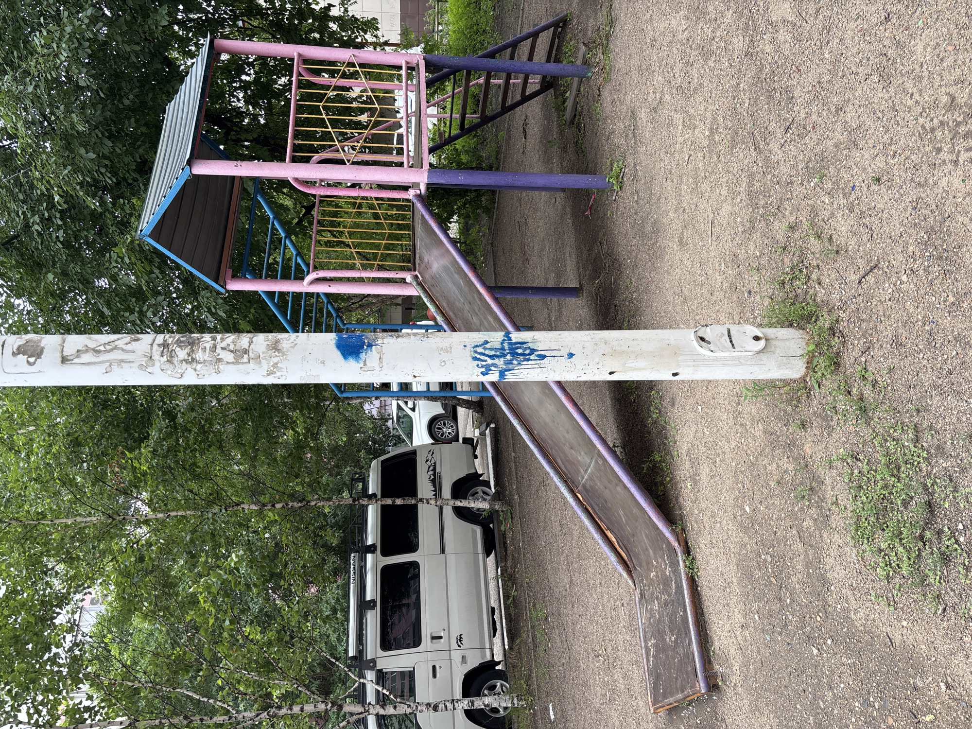 In Blagoveshchensk, Russia, on a cloudy day, a weathered playground slide and climbing structure stand on a dirt and gravel surface surrounded by trees. The slide, made of aged metal with purple trim, leads from a pink and yellow platform. A metal ladder with blue rungs ascends to the platform, which is topped by a corrugated metal roof with a blue edge. The structure is supported by purple posts. A white utility pole with graffiti, including blue spray paint, stands in the foreground, partially obscuring the slide and structure. To the left of the playground, a white Nissan van is parked, with roof racks and tinted windows. Another vehicle, a white SUV, is visible further in the background. The overall environment suggests an outdoor urban setting with signs of wear and tear on the playground equipment.