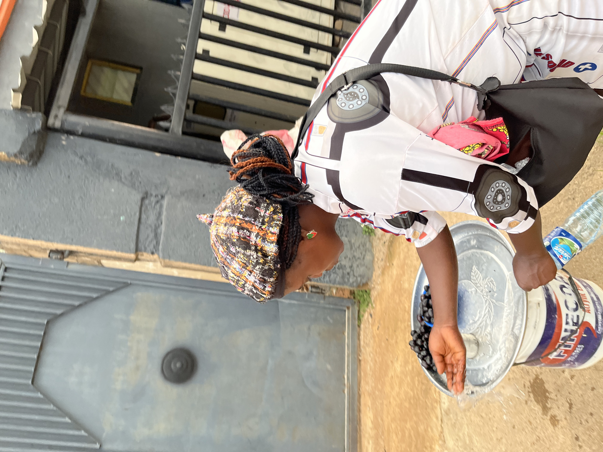 The image captures a person from a side-rear perspective, engaged in an outdoor activity, likely in Jos, Nigeria. The individual wears a white garment with prominent black, red, and blue geometric patterns, styled with a shoulder strap for a black bag. Their head is adorned with a multi-colored, textured hat covering braided hair, and a small, heart-shaped earring in green and red is visible on their earlobe.
The central activity involves the person bending over a silver metal basin that contains numerous dark, round objects, resembling grapes or berries. Water is actively being poured or splashing from the basin onto the brownish ground. One hand holds a clear plastic water bottle, labeled "Soeur WATER," suggesting it's being used to supply or manage the water. Adjacent to the basin is a large white plastic bucket with red and blue branding, displaying the text "FINECOAT ACRYLIC PAINTS."
The background on the left features a corrugated gray metal gate or door. In the upper-center, a dark window grill or railing is visible, with a rectangular framed object inside. The scene appears to take place during the day, with ample natural light, indicating an outdoor setting, possibly a street or a residential area.