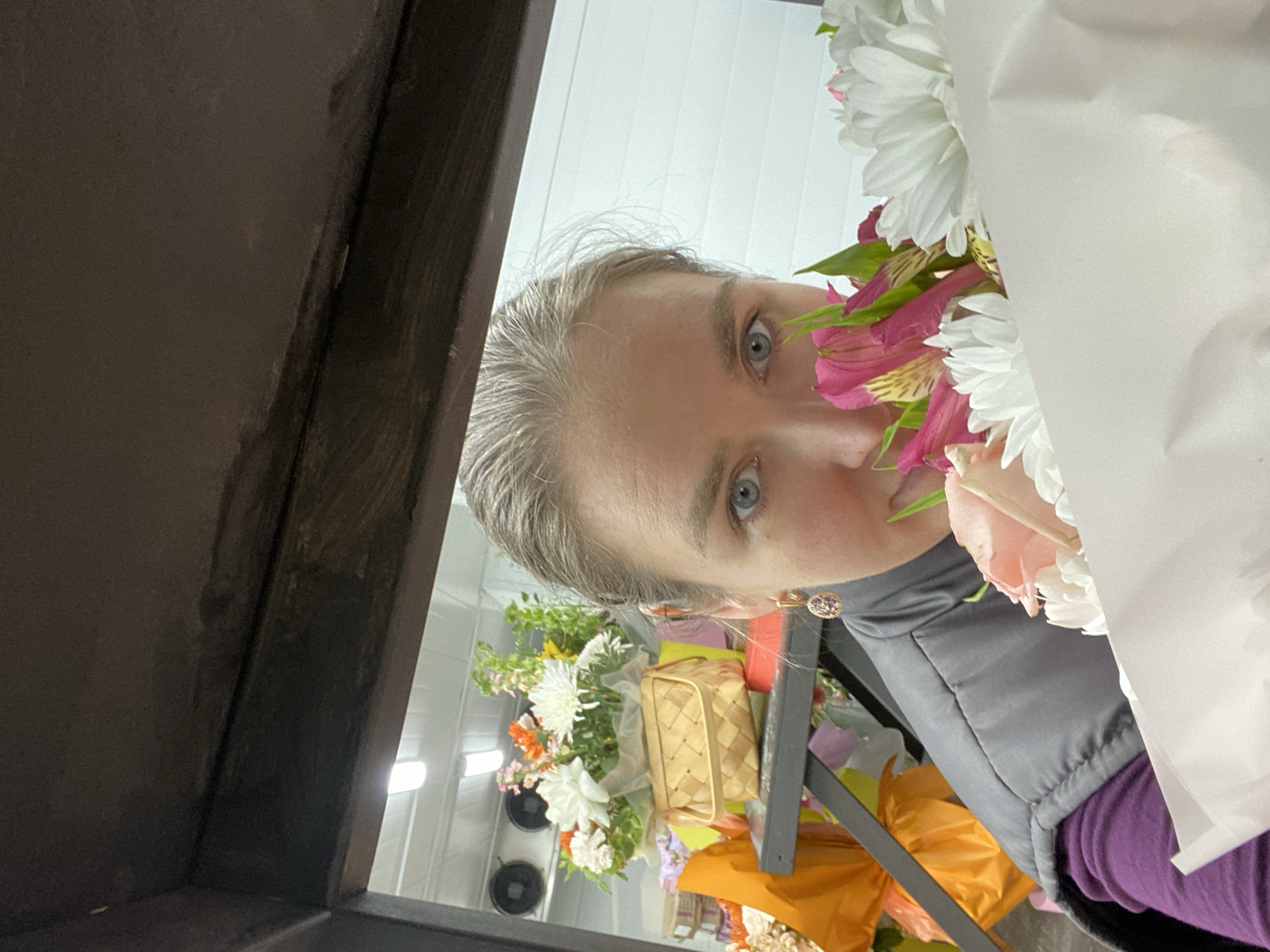 A pale-skinned person with gray hair and blue eyes partially obscures their face behind a bouquet of white and pink flowers. The person wears a gray vest over a purple shirt and a small, circular earring is visible. The scene appears to be inside a flower shop, with various other flower arrangements and a woven basket visible in the background. The location is potentially a flower shop in Yekaterinburg, Russia, although this detail is inferred and not explicitly visible in the image. The camera angle is unusual, possibly from inside a display case.