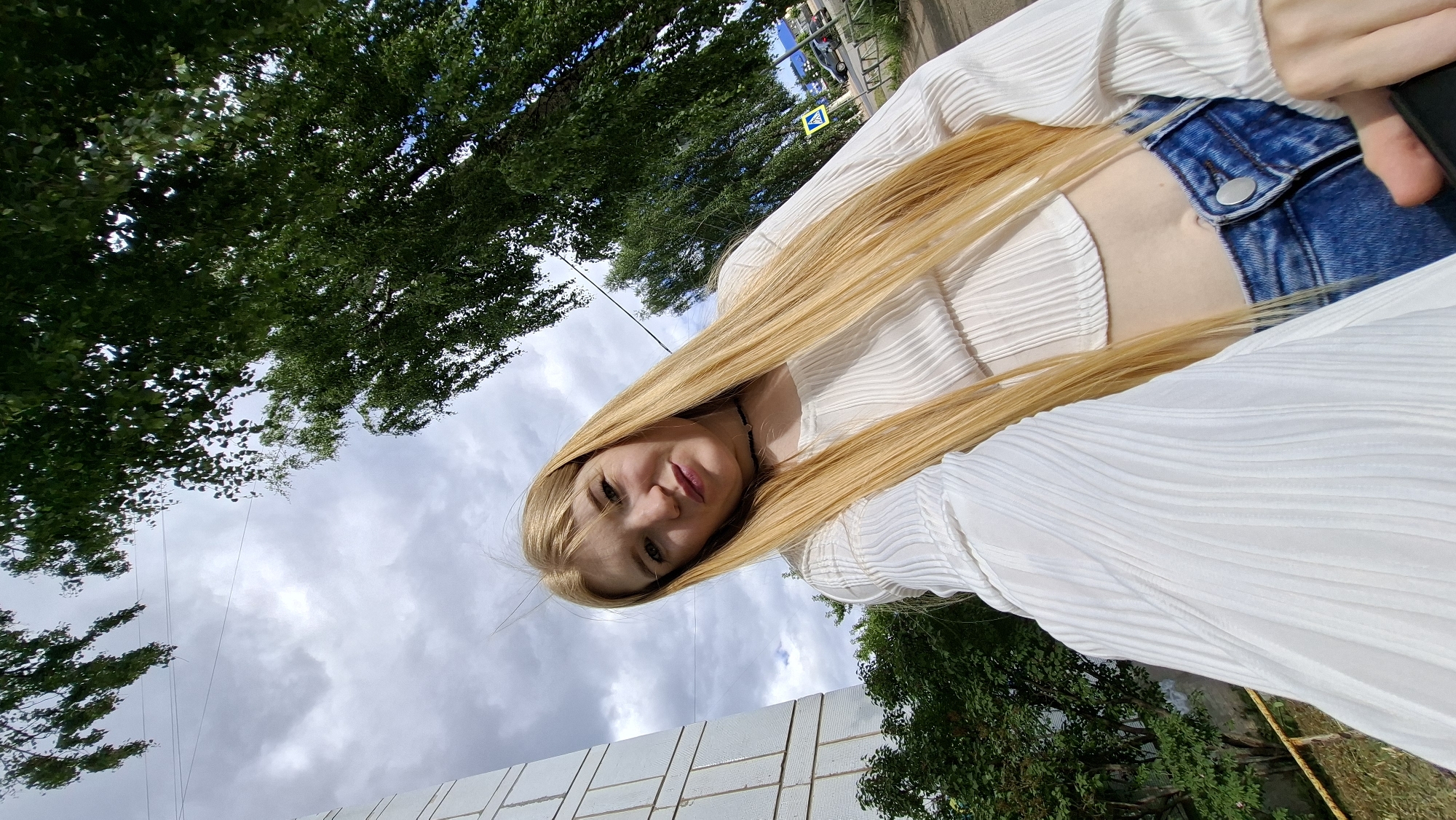 This is a slightly tilted, eye-level selfie taken outdoors on a cloudy day. The primary subject is a young woman with long, blonde hair and fair skin, wearing a white, pleated top and blue jeans. She is looking towards the camera with a slight smile. Her hair falls forward, partially obscuring her face and resting on her shoulders and chest. The background is a mix of lush green trees, a cloudy sky, and the corner of a light-colored, tiled building. In the far background, a street is visible with a car and a yellow traffic sign with a pedestrian symbol, suggesting an urban or suburban environment. The overall mood is casual and relaxed. The location is Chaikovsky, Russia.