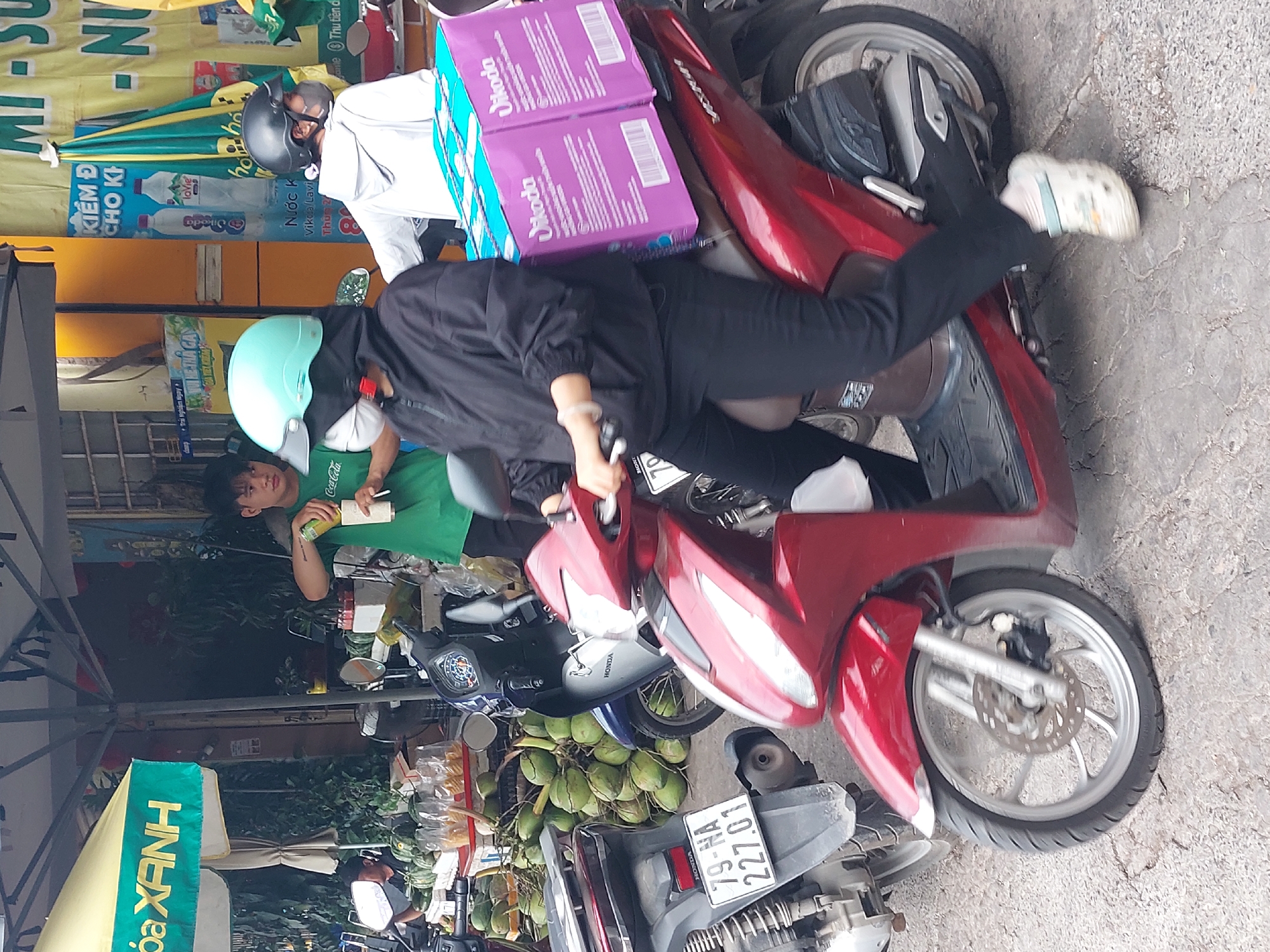 The image depicts a street scene in Nha Trang, Vietnam, with a focus on a young person on a red scooter carrying two large purple boxes labeled "Vikoda". The person is wearing a black hooded jacket, black pants, a light blue helmet, and a face mask. Another person wearing a white hoodie and a black helmet is visible on a separate scooter in the background. To the left, a man in a green shirt is serving drinks from a stall, with stacks of coconuts and other produce visible. The environment appears to be a market or roadside stall, with signs and awnings indicating a commercial area. The ground is paved with cracked concrete, and other motorbikes are parked nearby.