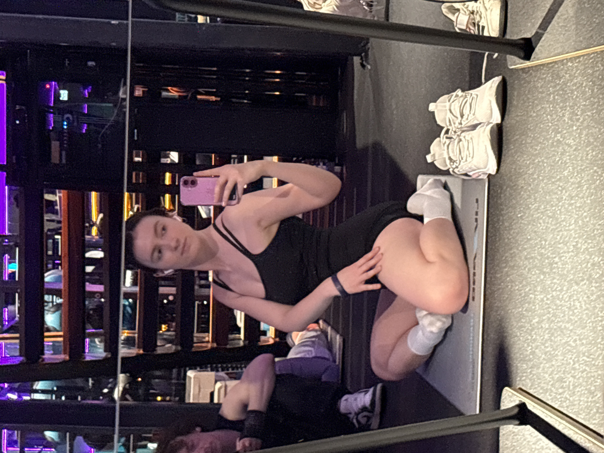 This mirror selfie, rotated clockwise in its original presentation, depicts an indoor scene likely within a modern gym or fitness studio in Kazan, Russia.
**Subjects:** The primary subject is a young woman, seen kneeling on a grey exercise mat, taking the picture with a pink smartphone held in her right hand. She has dark hair, fair skin, and a neutral expression as she looks directly into the mirror. She is wearing a black athletic top or bodysuit with thin straps and a dark wristband on her left arm. White socks are visible on her feet. In the lower left quadrant of the reflection, another person is partially visible, lying or sitting on the floor, dressed in dark attire and wearing white sneakers, suggesting another individual present in the facility.
**Objects:** Key objects include the pink smartphone used for the selfie, the grey exercise mat beneath the main subject, and multiple pairs of athletic shoes. A pair of white sneakers with white laces is visible on the floor to the right of the mat, and another pair of light-colored shoes is further in the background.
**Scene and Setting:** The background, visible through the mirror's reflection, consists of dark structural elements, possibly gym equipment or internal architecture. The lighting is predominantly artificial, with distinct purple and orange hues casting mood lighting across the space, indicative of an evening or night-time setting. The flooring is a textured grey, common in fitness environments.
**Activity:** The central activity is the woman taking a mirror selfie.
**Notable Details:** The colored artificial lighting creates a modern and vibrant atmosphere. The image captures the depth of the room through the mirror's reflection. There is no visible text in the image.