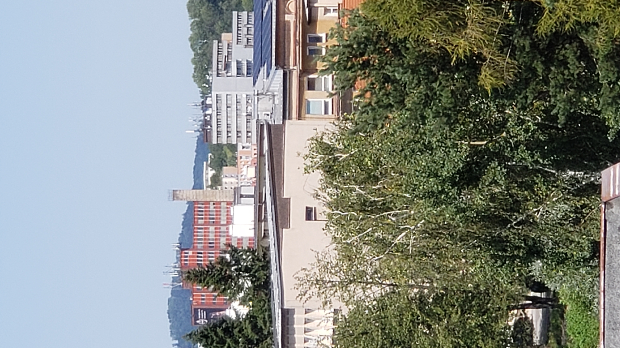 The image captures an urban landscape with a clear blue sky on a bright day. In the foreground, lush green trees, including a prominent birch with white bark, dominate the scene. Behind the trees, several buildings are visible.
To the left, a tall, red brick building with numerous windows is a focal point. It features a distinctive industrial chimney or tower adjacent to it. Further to the right, a long, grey, multi-story building with many windows and what appear to be communication antennas on its roof is visible. In the middle ground, a lighter-colored building with a flat roof and some architectural detailing is partially obscured by the trees. Several other buildings of varying heights and architectural styles, including one with solar panels on its roof, are scattered throughout the cityscape. The background shows rolling hills covered in greenery, suggesting the city is situated in a relatively natural setting. The overall impression is one of a developed urban area surrounded by nature.
This is a view of the area around the former textile factory "Zornica" in Prešov, Slovakia. The red building in the background is likely the main factory building, and the tall structure next to it is an old chimney or ventilation tower. The grey building with antennas is also likely part of the industrial or commercial area.