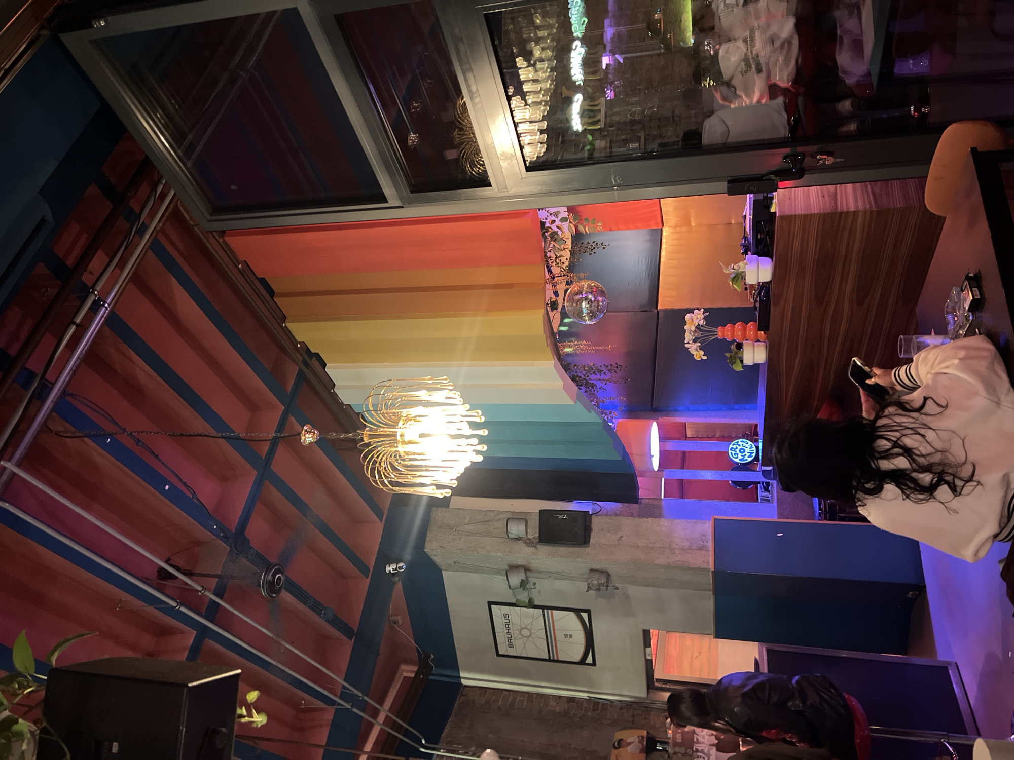 The image depicts an indoor scene likely in a restaurant or bar in Belgrade, Serbia, characterized by unique interior design. The space is divided by structural elements; on one side is the ceiling constructed of shipping containers, painted with red and blue stripes. A decorative chandelier hangs from the ceiling. The other side of the interior features a rainbow-striped wall leading to a decorated area with plants and a disco ball. The wall also has a floral decoration.
A skylight runs along the ceiling, reflecting the lights of the city. A table is placed to the side of the skylight, and a person with long, dark hair wearing a white jacket is seated at the table, looking at a phone. Another person in a dark jacket is visible in the background, next to a blue panel. There is what appears to be a Bauhaus design wall hanging, light fixtures, and other structural support elements visible on the wall. The overall environment is dimly lit with colorful lighting.