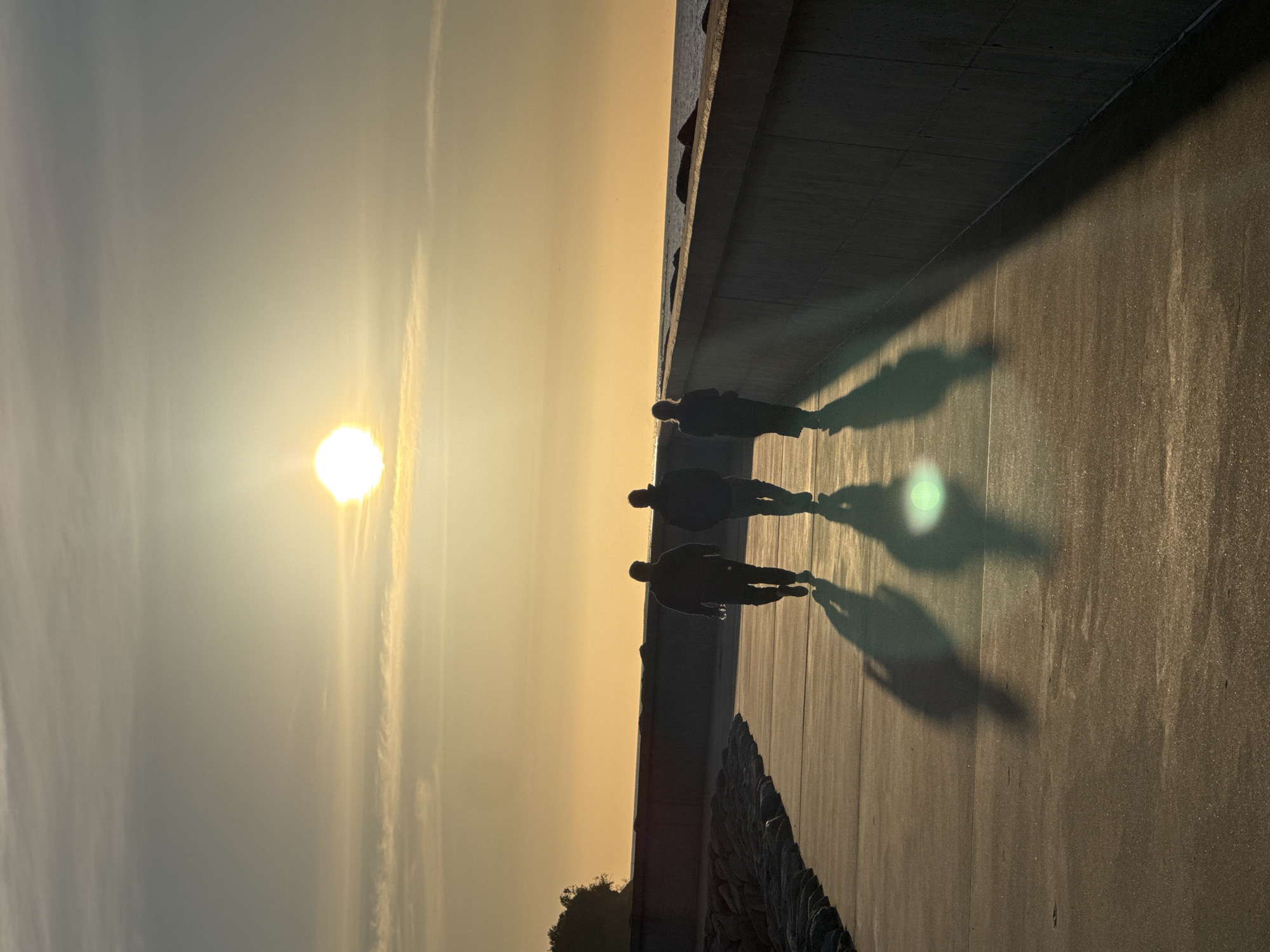 The image shows three silhouetted figures walking along a paved promenade next to a concrete barrier wall. The sun is low in the sky, casting long shadows from the figures onto the promenade. The promenade appears to be located in an urban setting, possibly a coastal area, given the presence of the sea and a rocky embankment to the left. The sky is a warm golden-orange hue, indicating either sunrise or sunset. The overall atmosphere is serene and peaceful. The scene is consistent with the Pärnu Promenade in Tallinn, Estonia, a popular waterfront area known for its scenic views and walking paths.