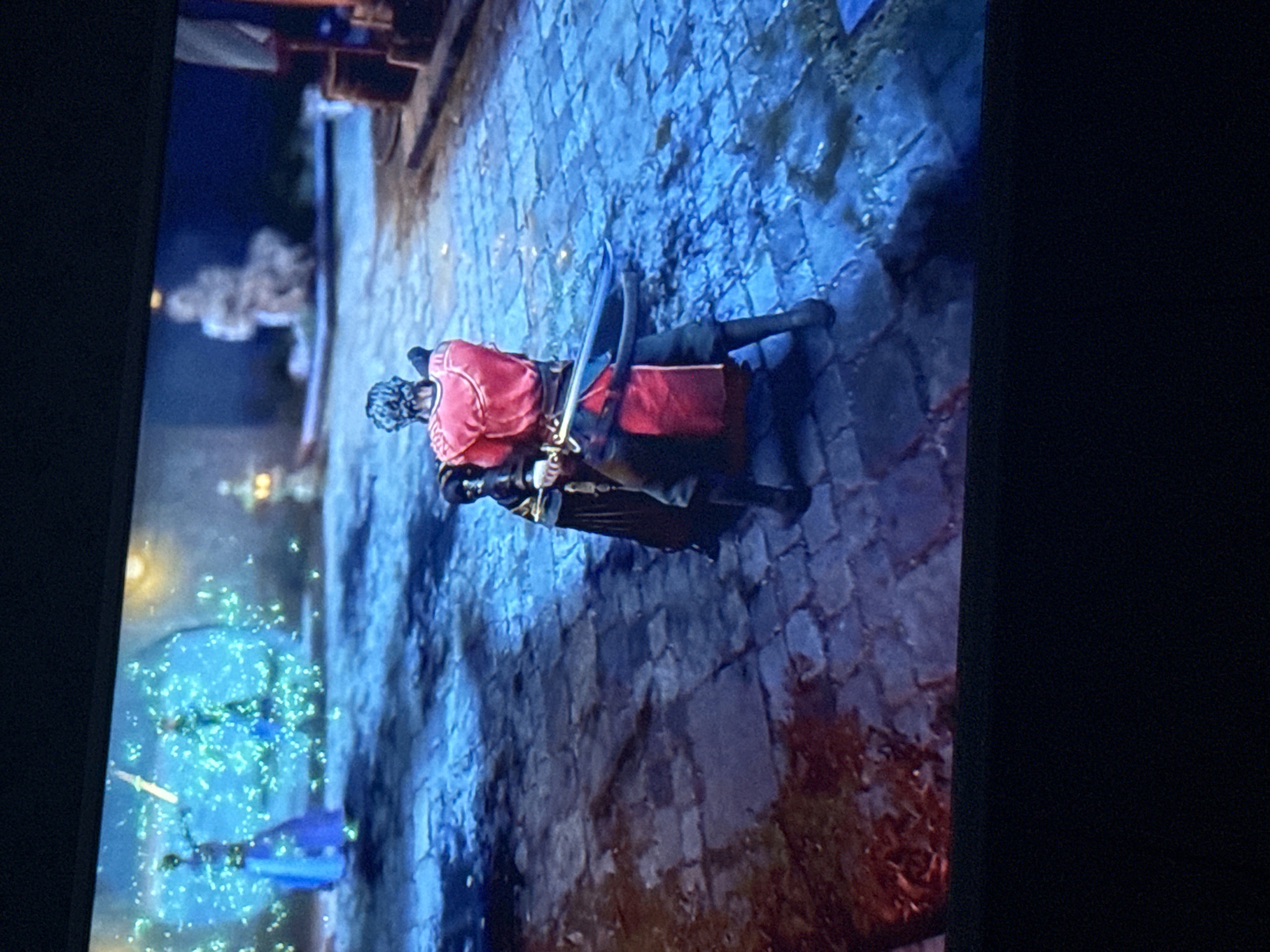 A television screen displays a video game scene set on a cobblestone street at night. Two figures are the primary focus: One, wearing a red coat, stands over the other, who is clad in dark clothing. The figure in red holds a sword. The setting appears to be an urban environment, possibly a historical city. The background features indistinct buildings and a bright light source, creating an atmosphere that blends elements of fantasy or historical fiction. The game's graphics show detailed textures and lighting effects, enhancing the visual realism of the scene. It is not clear where this image was taken, but it cannot be confirmed that it was taken in Mytishchi, Russia.