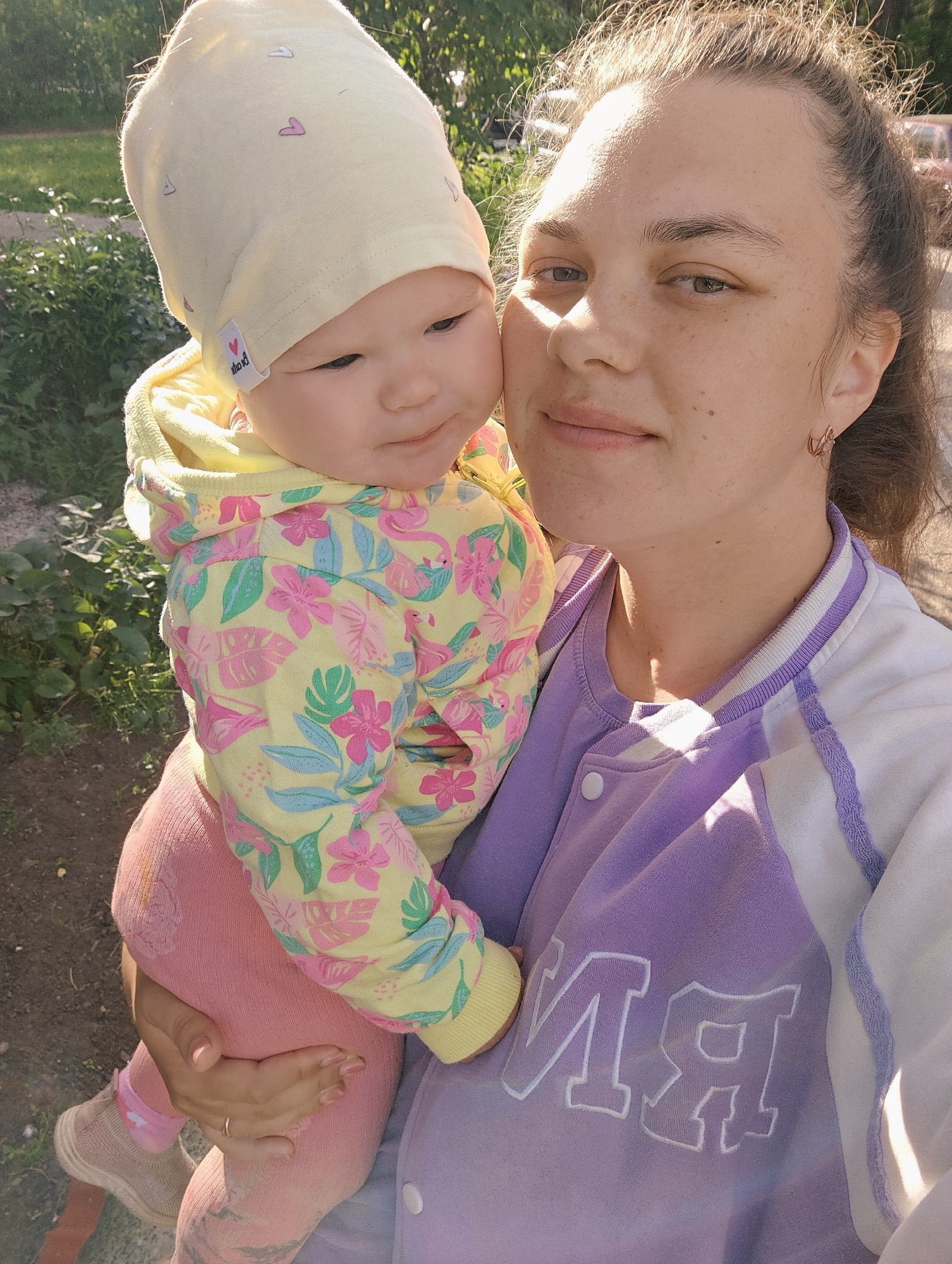 A woman is holding a baby in an outdoor setting, possibly in a city like Chaikovsky, Russia. The woman, who appears to be in her late 20s to early 30s, has her hair pulled back and is wearing a lavender and white jacket. She has light skin and minimal visible makeup. The baby, seemingly under a year old, is wearing a light yellow hat and a brightly patterned jacket featuring floral and flamingo designs. The baby is also wearing pink leggings and sneakers. The background suggests a park or green space with trees and vegetation. The image is taken in natural light, with a soft, warm glow indicating it might be during the late afternoon or early evening.
