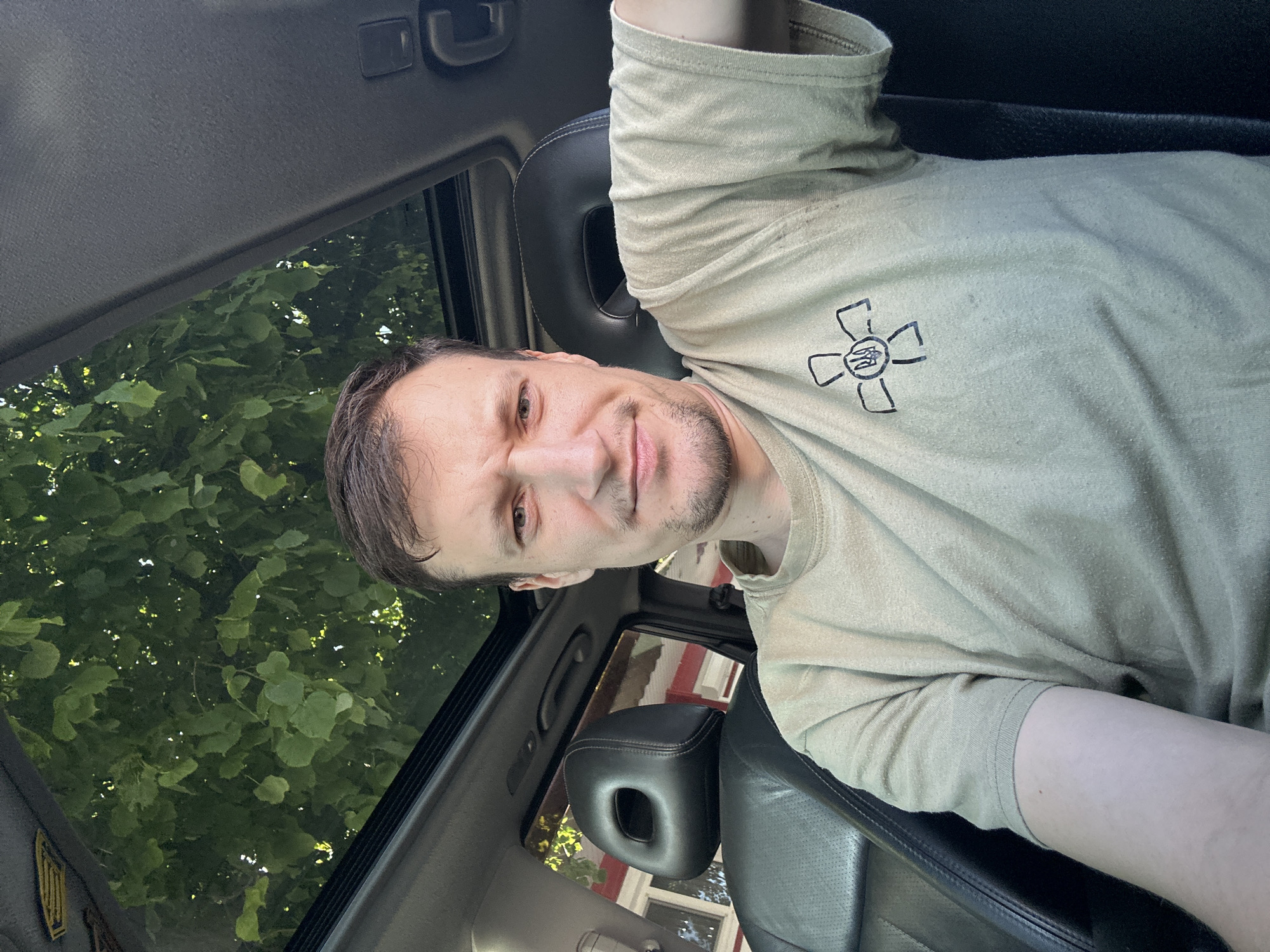 A selfie is taken by a man sitting in the driver's seat of a car. He is wearing a light brown t-shirt with a black emblem of a cross with a trident in the center. The car has a sunroof open, revealing green tree leaves and branches above. The interior of the car is visible, including black leather seats and part of the dashboard. In the background, through the car window, a building with red and white sections and windows is visible. The man has short brown hair, a light beard, and is looking directly at the camera with a slightly furrowed brow. The image appears to have been taken in Kamianets-Podilskyi, Ukraine.