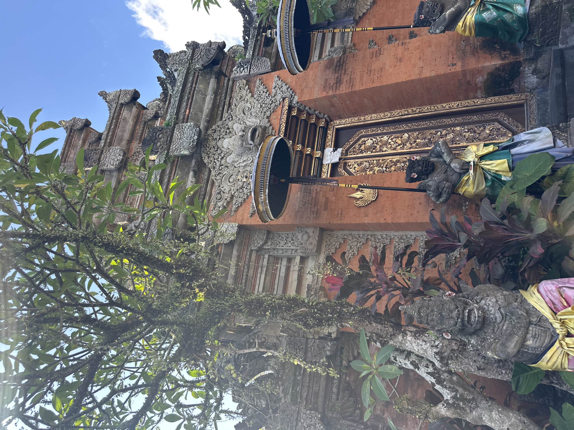 The image captures an exterior view of a Balinese temple entrance, likely located in the Ubud District, Indonesia. The focal point is a large, intricately carved brick temple gate, adorned with detailed stone sculptures of mythical creatures and deities. Two ornate, dark umbrellas stand on either side of the entrance, their golden embellishments catching the light. In front of the gate, two stone guardian statues, dressed in traditional Balinese sarongs, are positioned to protect the entrance. Lush green foliage and trees surround the temple, with one particularly large, moss-covered tree to the left, its branches reaching towards the sky. The sky above is a clear blue with a few white clouds, indicating a sunny day. The scene is tranquil and evokes a sense of cultural and spiritual significance.
