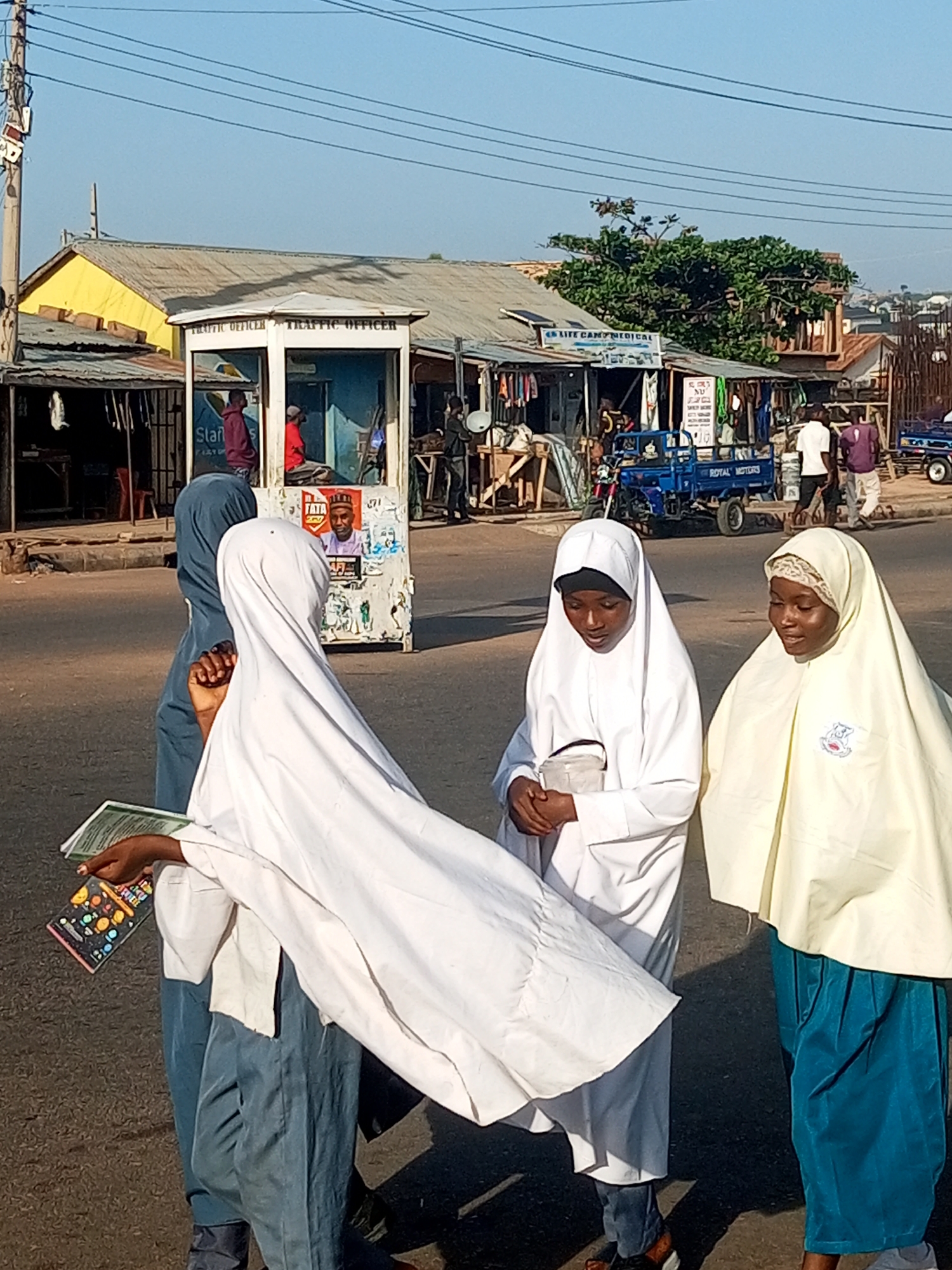 In an outdoor urban setting, four young individuals, three of whom are wearing hijabs, are visible in the foreground. Two of the individuals in hijabs are looking down and to their right, while the third individual in a hijab is looking to her left. The fourth individual, who is not wearing a hijab, is turned away from the camera and appears to be holding a book. In the background, a traffic booth with the words "TRAFFIC OFFICER" is situated on the side of a street. Several structures, possibly shops or stalls, are lined up behind the traffic booth. A blue vehicle with a trailer is parked nearby. Power lines are suspended overhead against a clear sky. The scene appears to be during daylight hours.