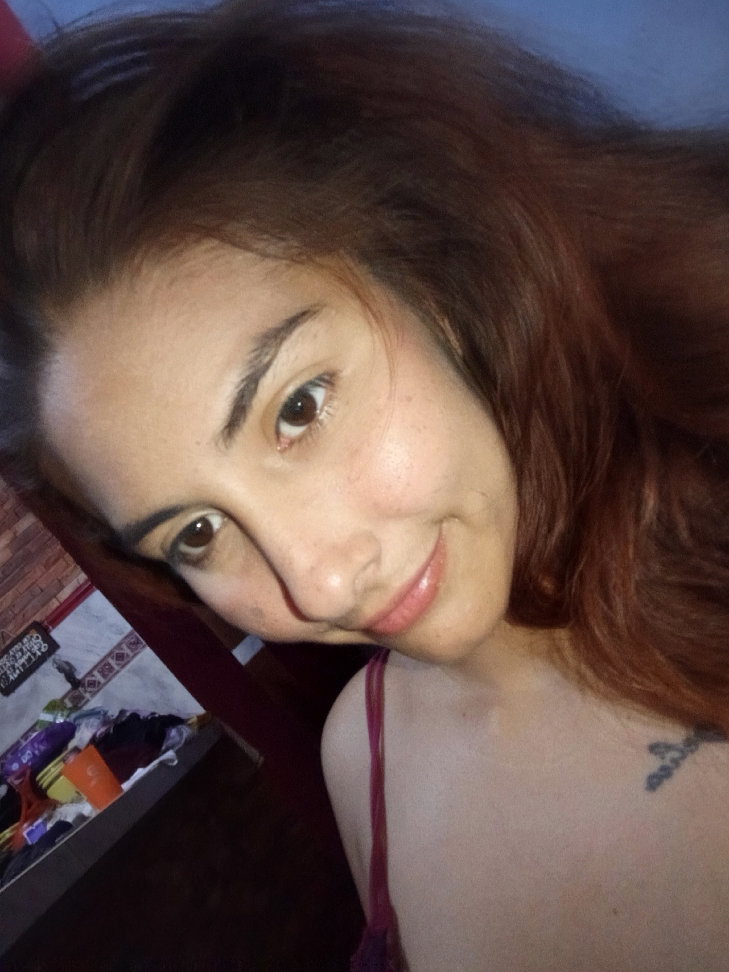 A selfie of a woman with medium-length auburn hair, light skin, and brown eyes. She is wearing a red tank top. A tattoo is visible on her chest. The background includes a cluttered surface with various items and a wall. The location is indicated as Juan Griego, Venezuela, but there are no identifiable landmarks or geographical features to confirm this within the image itself.