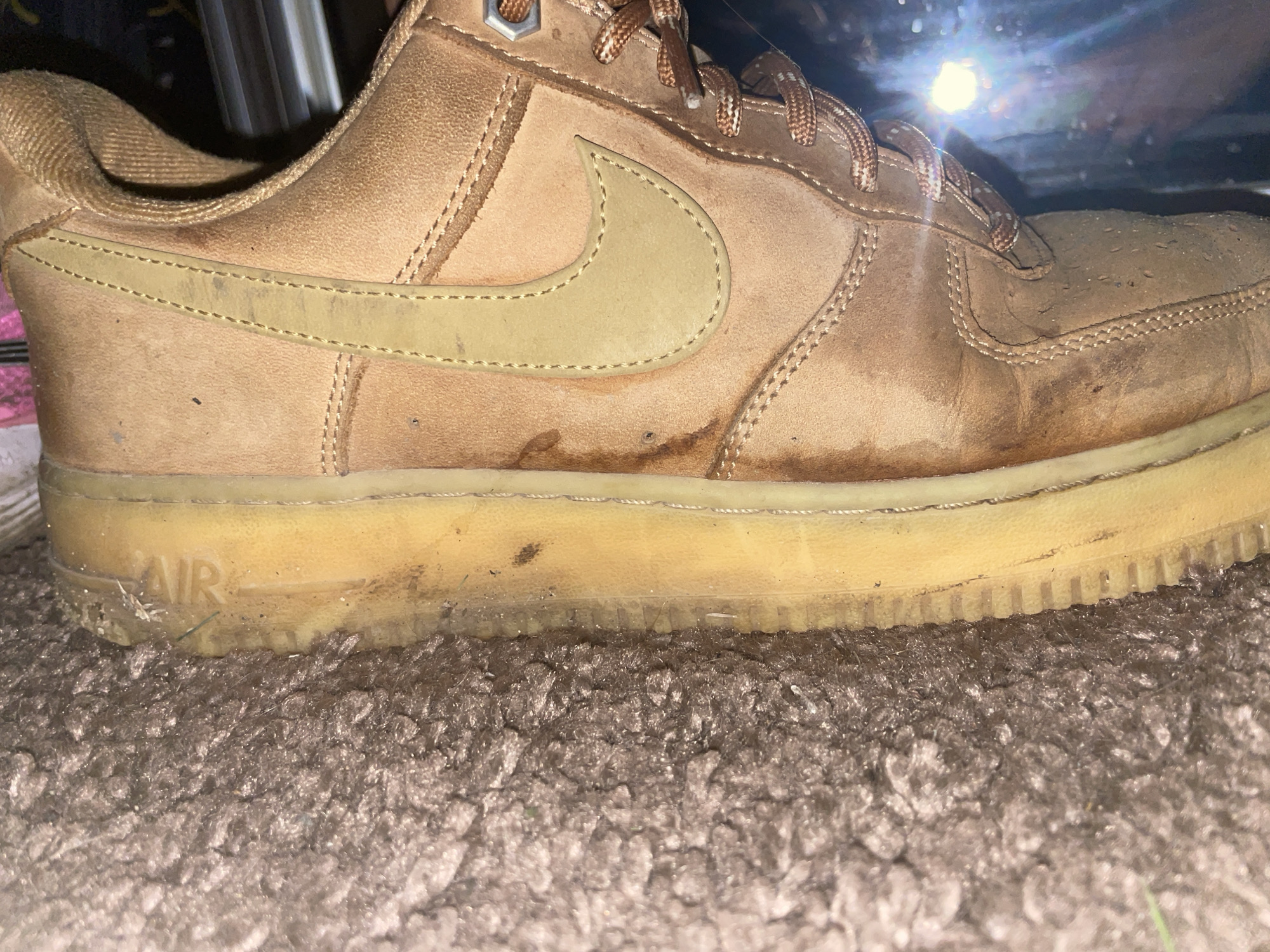 A close-up, side view of a tan Nike Air Force 1 sneaker with a visible "AIR" logo on the sole. The sneaker appears worn and dirty, with water stains and scuffs on the leather and suede upper. The laces are also tan. The shoe is resting on a textured, grey carpet. The background is dark and out of focus, with a bright light source visible at the top right, creating a glare. The location is Unknown, Unknown.