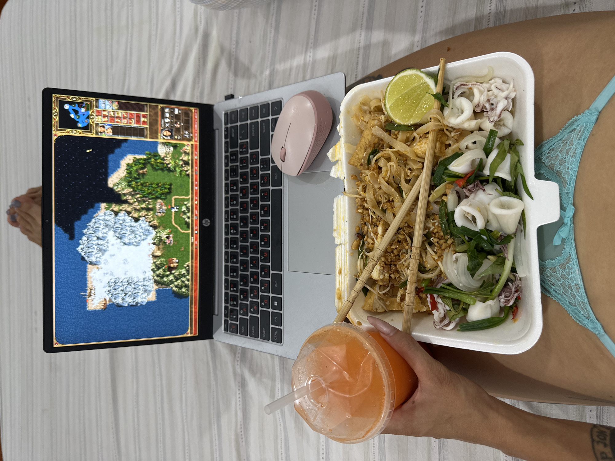 Here is a detailed description of the image:
**Overall Scene:** The image presents an overhead view of a person engaged in leisure activities on what appears to be a bed or similar soft surface. The focus is on a laptop, a meal, and a beverage, indicating a relaxed setting.
**Objects:**
* **Laptop:** A laptop is positioned centrally, its screen displaying a video game.
* **Mouse:** A pink wireless computer mouse.
* **Meal:** A takeout container of what looks like Pad Thai with additional squid, lime and vegetables is placed beside the laptop.
* **Drink:** A clear plastic cup filled with an orange liquid, presumably a juice or beverage, held by the hand of the person.
* **Chopsticks:** Wooden chopsticks are inserted within the Pad Thai.
**People/Anatomy:**
* A hand holds the drink, and a foot is visible on the side of the computer.
* The person is wearing underwear that is turquoise in color.
**Actions/Interactions:**
* The person is likely playing the game on the laptop while simultaneously enjoying their meal and drink.
* The placement of the objects suggests an intent to multitask, possibly enjoying a meal during a break.
**Environment:**
* The background appears to be a bed with striped bedding, giving a casual and private atmosphere.
**Location:**
The location in the city of Mũi Né, Vietnam cannot be identified from the image.