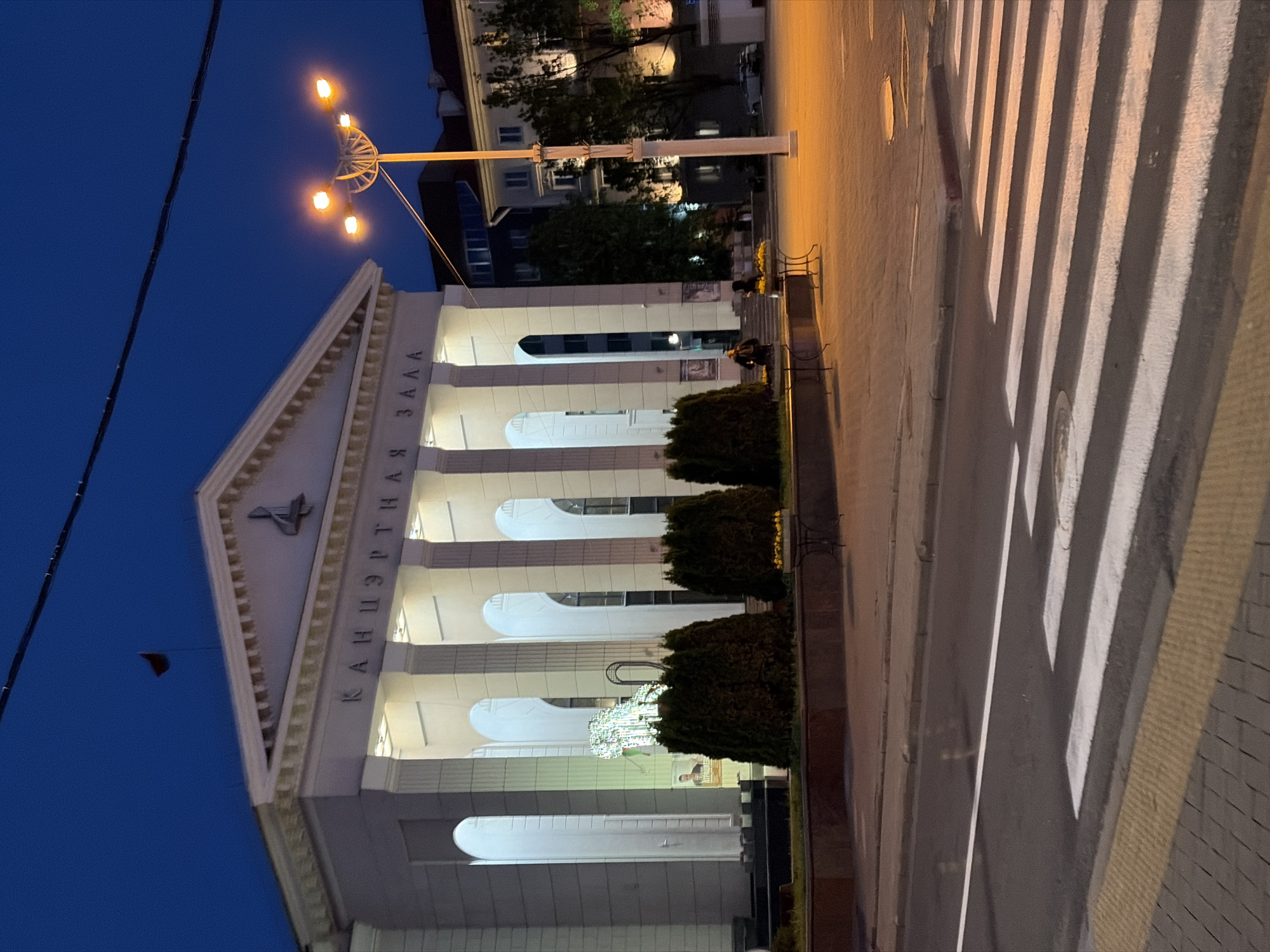 The image shows the Concert Hall "Minsk" in Minsk, Belarus, at dusk. The building is a classical structure with white columns and illuminated arches. The inscription "КАНЦЭРТНАЯ ЗАЛА" (Concert Hall) is visible on the facade. A tall street lamp with multiple lights stands nearby. In front of the building, there are several small, neatly trimmed bushes. A zebra crossing is visible on the street adjacent to the building. There are two people on the steps of the concert hall. The sky is a deep blue, indicating that it is twilight.