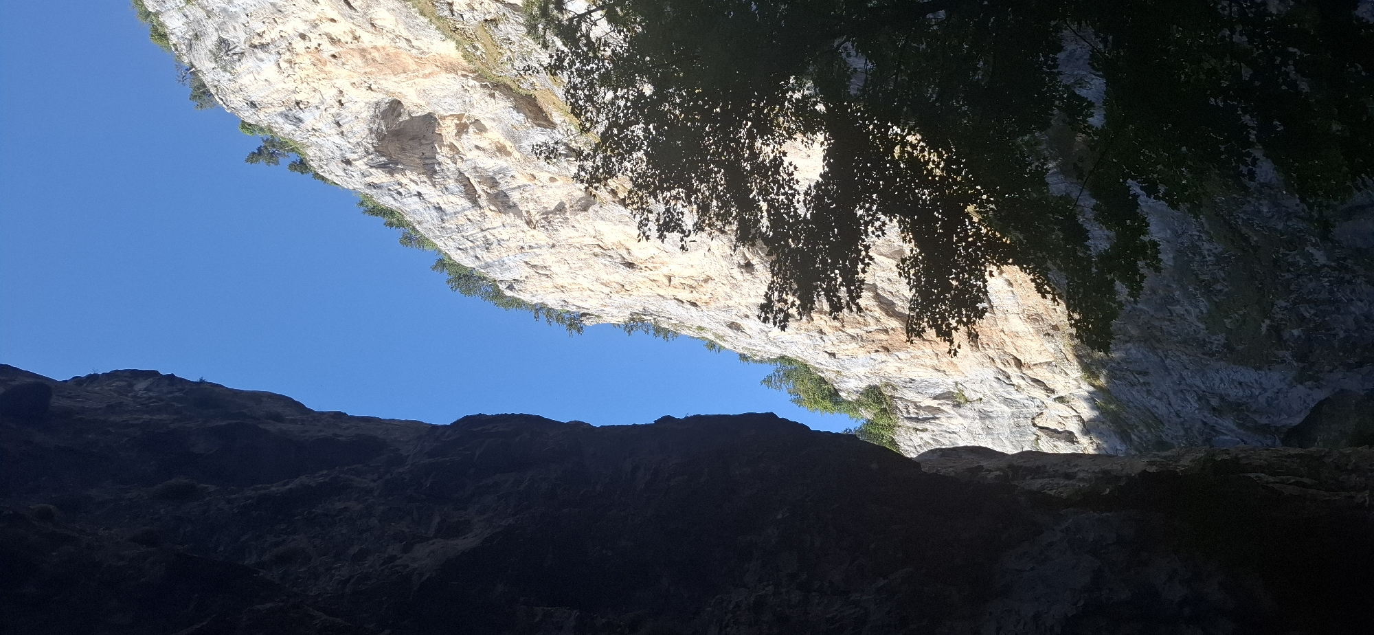 The image shows a view from a narrow gorge, looking upwards towards the sky. On the left, a dark, imposing cliff face dominates the foreground, casting a deep shadow. On the right, a lighter, sunlit cliff rises steeply, revealing layers of rock and small patches of vegetation clinging to its surface. The sky above is a clear, vibrant blue. The scene appears to be a natural landscape, likely within a mountainous region. The location is identified as the Trigrad Gorge in Trigrad, Bulgaria, a place known for its dramatic karst formations and caves. There are no people or animals visible in the image. The overall impression is one of natural grandeur and rugged beauty.