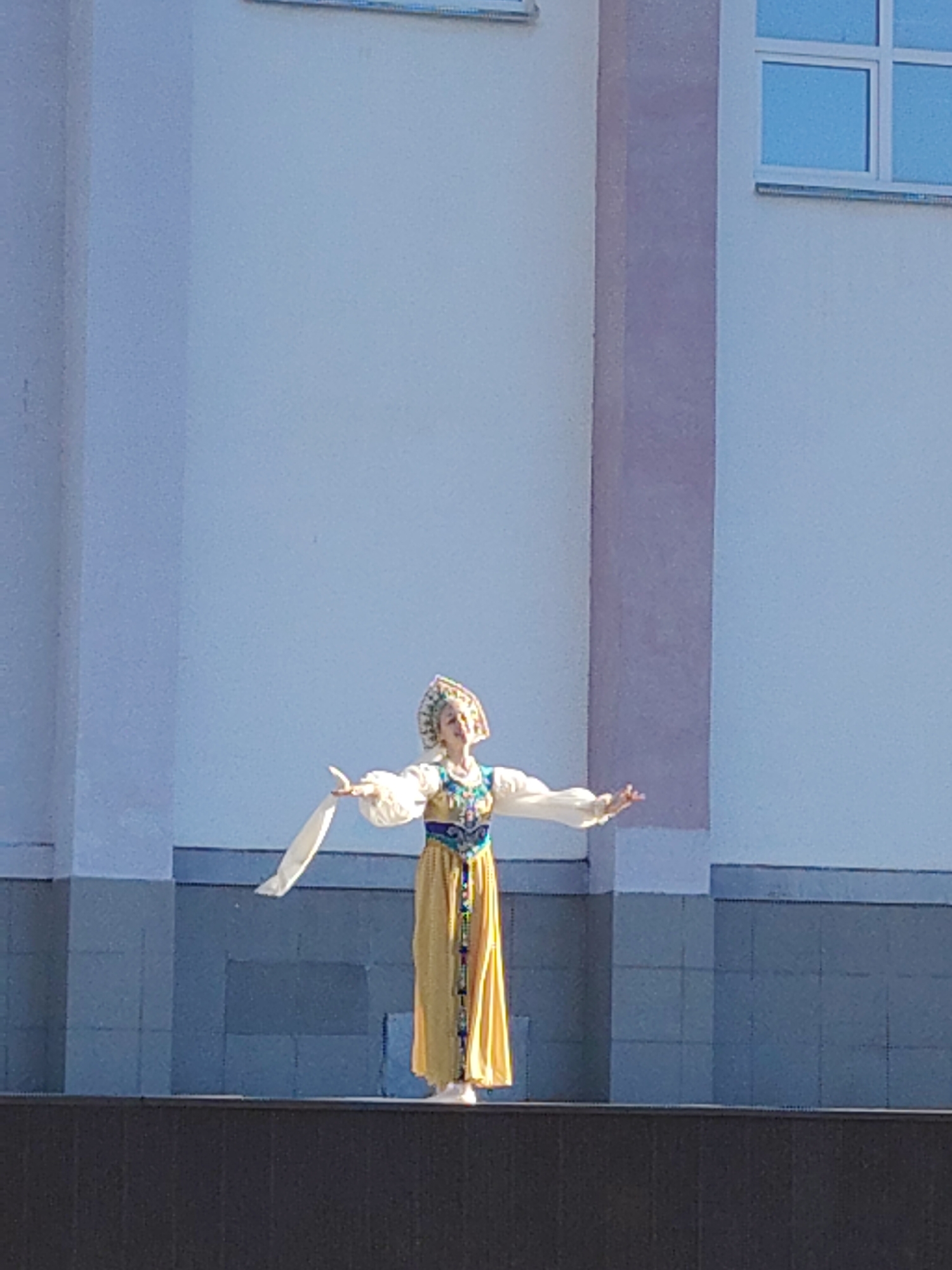 A woman in traditional Russian dress stands on a platform outdoors. Her attire consists of a long yellow dress, a white blouse with billowing sleeves, a decorative vest, and a traditional headdress. She is holding a white scarf in one hand and extending her other arm to the side. The background features a building with a white facade and grey accents, including a window with blue glass. The location is the city of Unknown, Unknown.