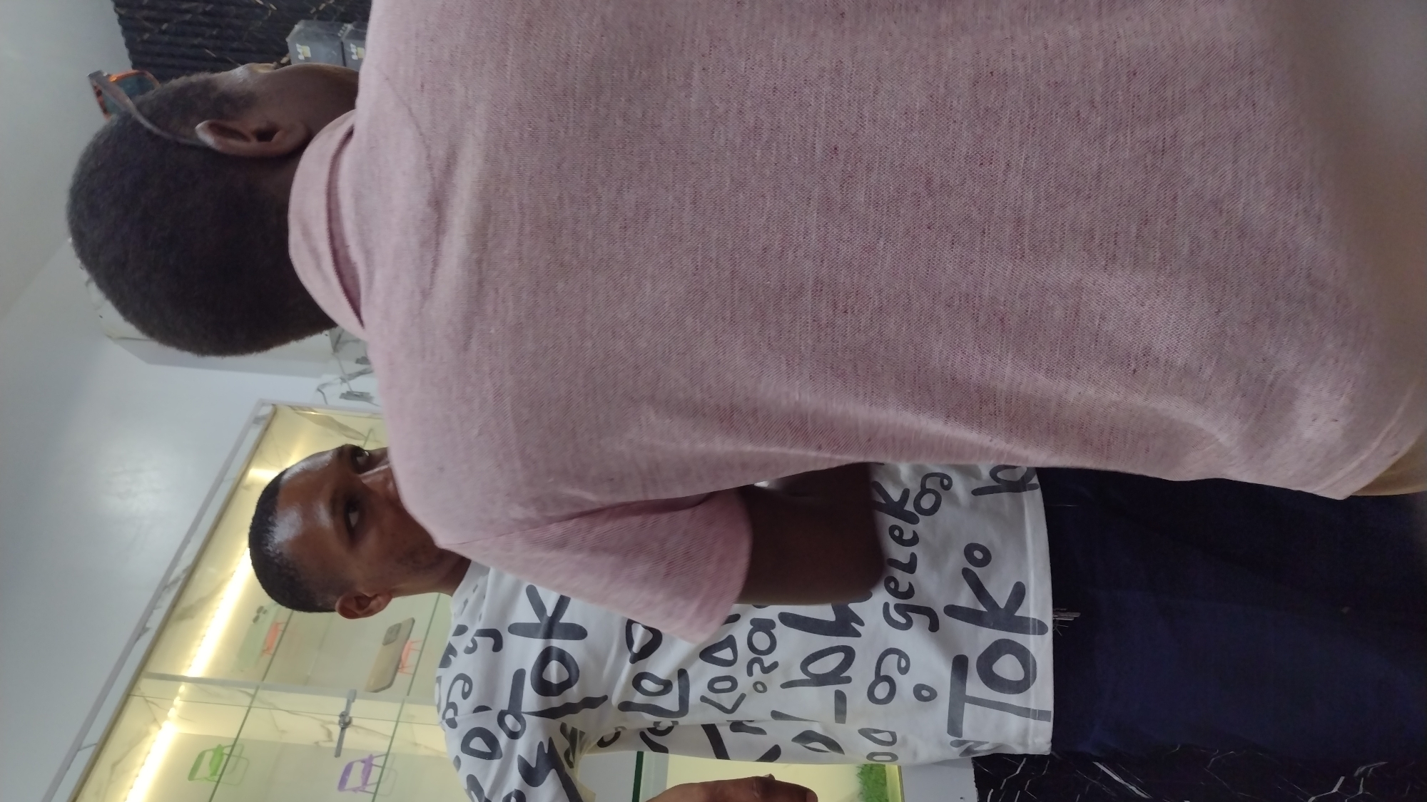 Two men are visible indoors. One man is closer to the camera and is wearing a pink short-sleeved shirt. The other man is wearing a white T-shirt with "Tok" and other indecipherable text printed on it. They seem to be in an optician's shop, with a display case of eyeglasses and other items visible in the background. The location is Unknown, Unknown.