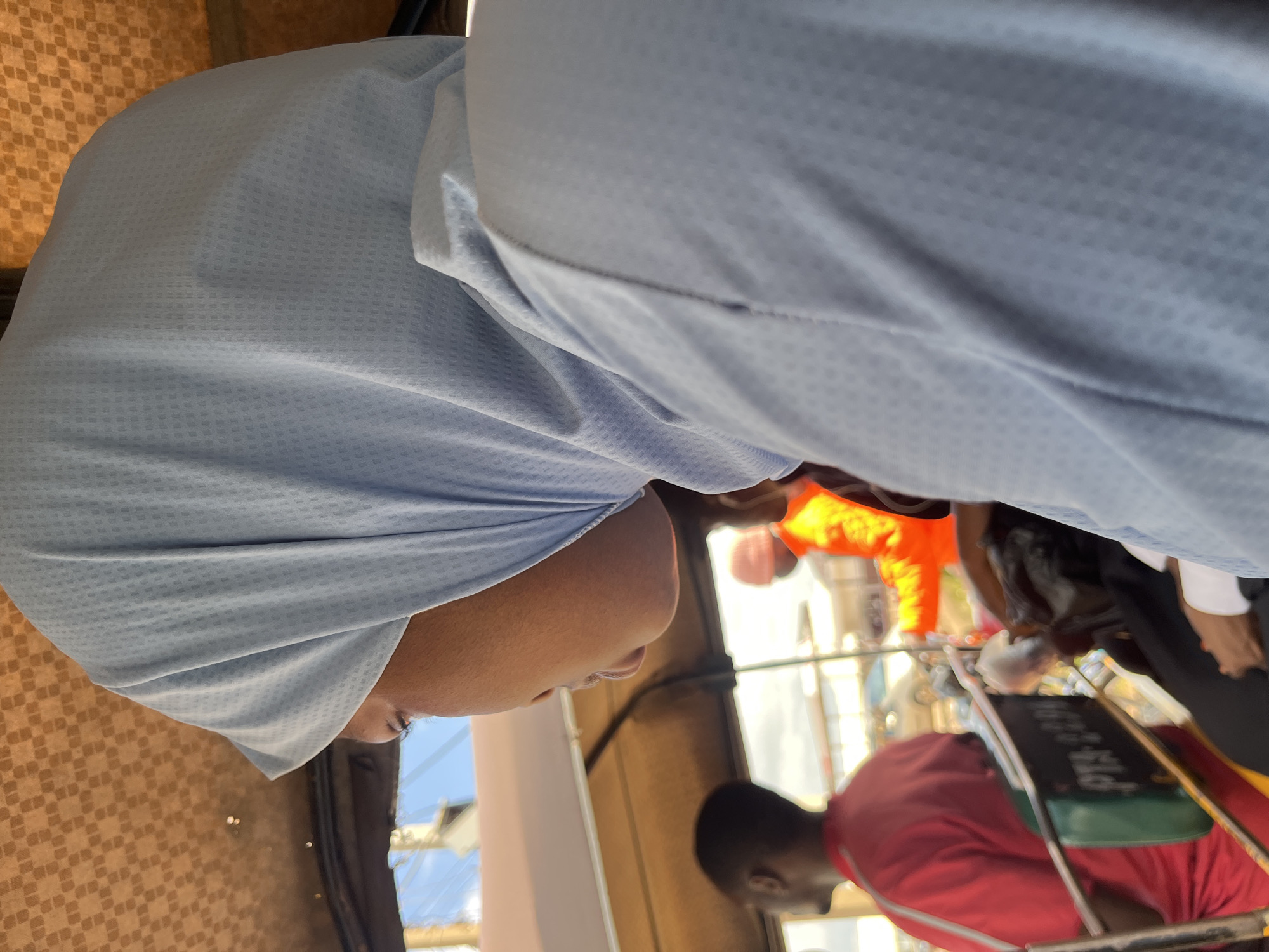 A headshot of a person wearing a light blue hijab in what appears to be a tuk-tuk. The person is looking to the left. In the background, another person in a red shirt is visible, and further back, someone in an orange jacket. The interior of the tuk-tuk has a patterned brown ceiling. The scene is outdoors with glimpses of the sky and structures in the distance. The text on a plate inside the tuk-tuk is partially visible.