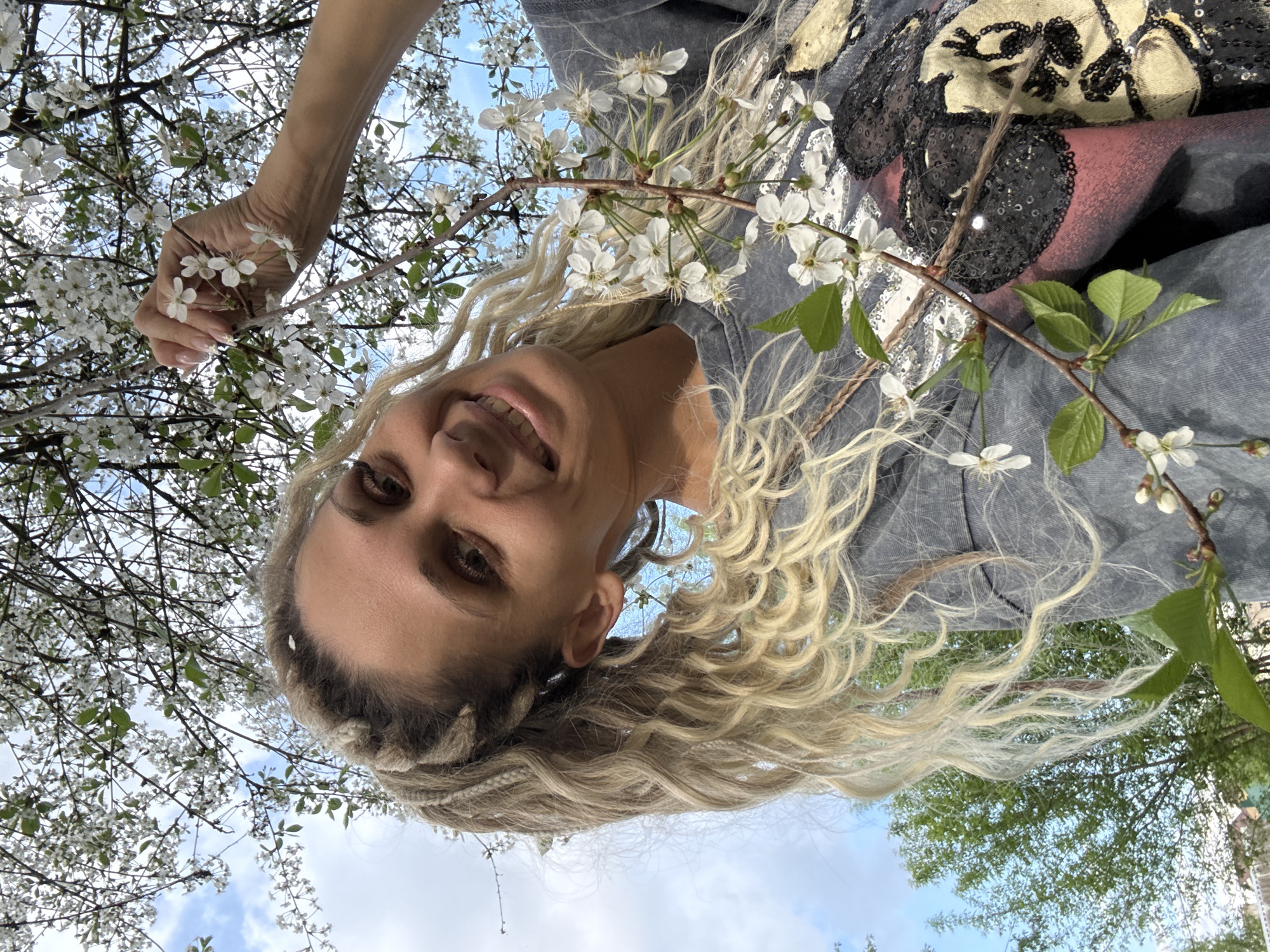 A woman with blonde hair is smiling at the camera. She is holding a branch of white flowers. The background is a blue sky and trees. The image was taken in Rusanovo, Russia.