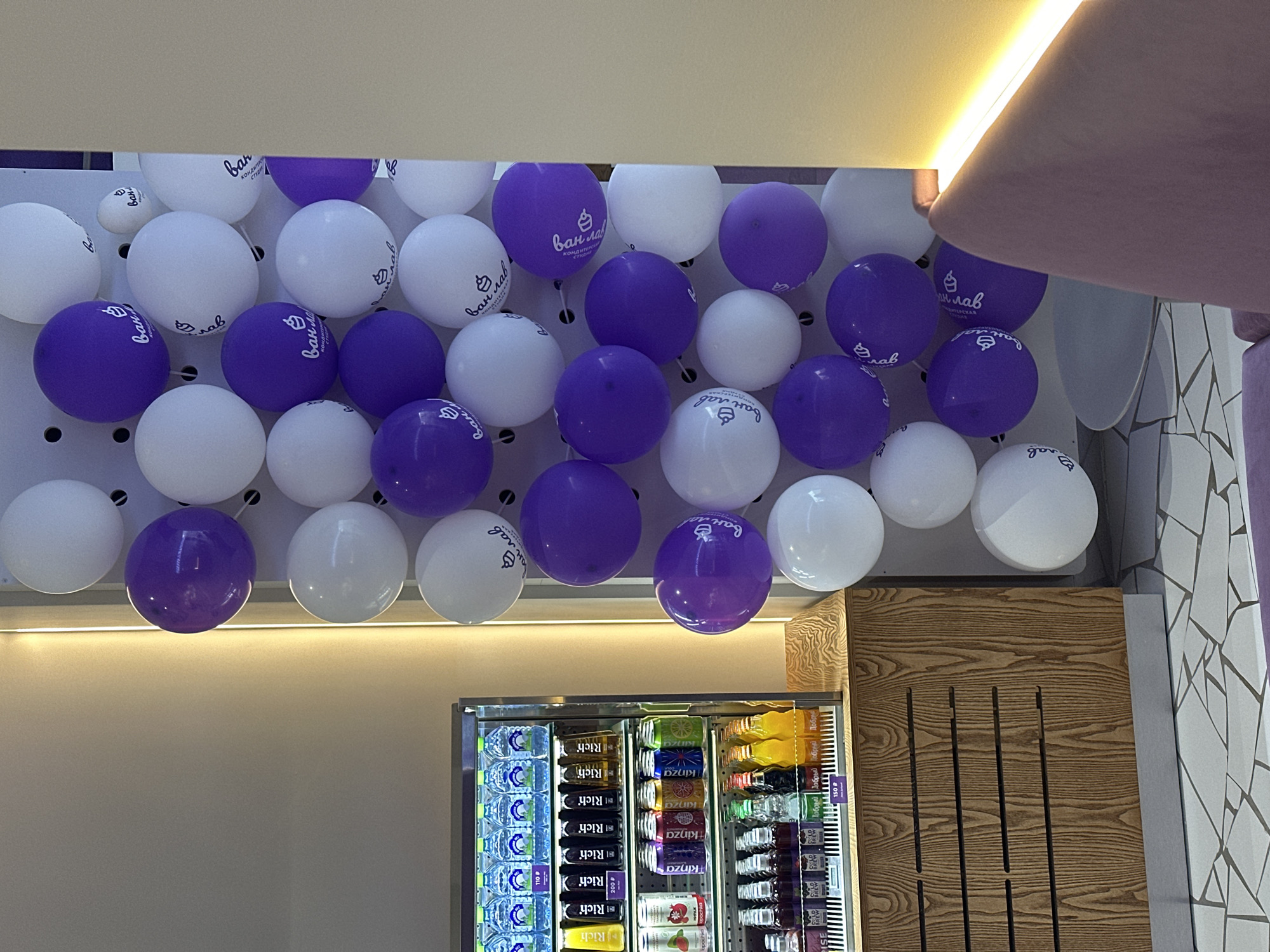 The image shows an interior space decorated with white and purple balloons that have the logo of "van-lav Konditerskaya Studiya" printed on them, suggesting it's a celebratory or promotional setting. Below the balloon decoration is a refrigerated display case stocked with bottled and canned beverages, including water, Rich juices, and Kinza drinks. To the right of the refrigerator is a wooden panel with vertical slats, and adjacent to that is a mosaic-tiled wall. The location is indicated by the text on the balloons, "van-lav Konditerskaya Studiya," which is a кондитерская студия (confectionery studio) in Kazan, Russia. The presence of Cyrillic text on the beverage labels confirms this location. The overall scene appears to be the interior of a cafe or shop related to "van-lav."
