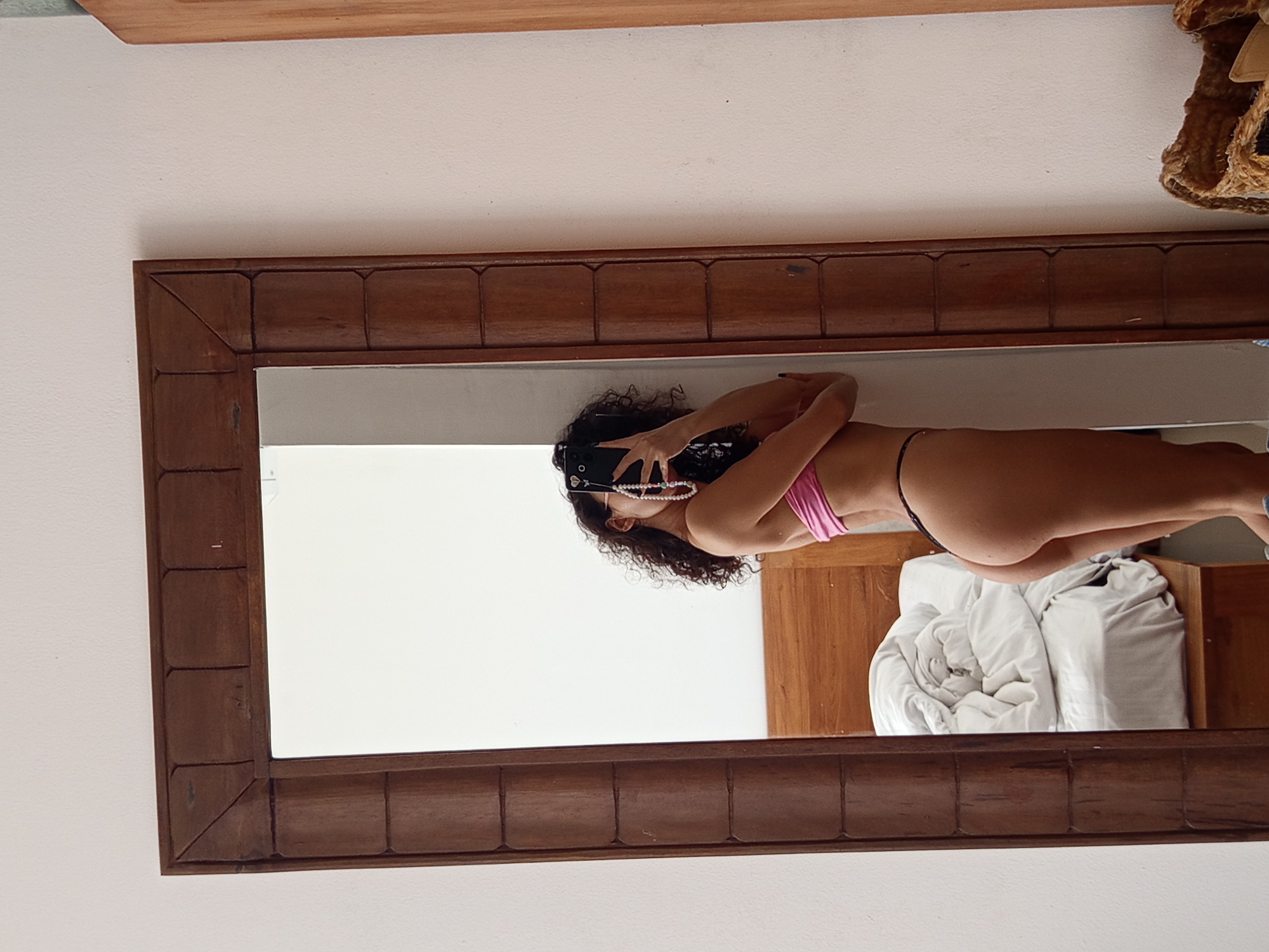 A woman is visible in the reflection of a wooden-framed mirror, likely a bathroom mirror. She is wearing a pink top and a black thong. Her hair is dark and curly, and she is wearing a pearl necklace. The woman appears to be taking a selfie, holding her phone in front of her face. The background of the reflection shows a wooden cabinet and a white comforter on a bed. The mirror is hanging on a white wall. The location is potentially in North Kuta, Indonesia.