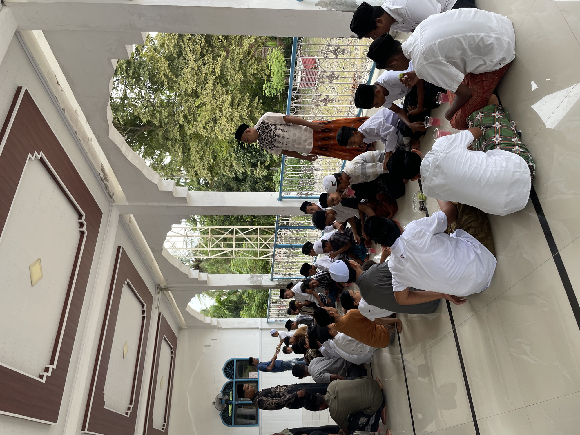 The image depicts an outdoor gathering of approximately 30-40 individuals, predominantly young males, seated on a tiled floor. They are dressed in traditional Islamic attire, including peci (caps) and sarongs. The setting appears to be a covered patio or veranda of a building, with white columns and a decorative ceiling visible. Through an open railing, lush green trees and foliage are visible, suggesting a tropical environment. Some individuals are interacting, with some appearing to be eating or receiving food. One man in the foreground is wearing a white shirt and patterned sarong, while another man in the background stands and appears to be addressing the group. The floor is light-colored tile with dark grout lines. The overall atmosphere suggests a communal event or meal. The location is identified as Trieng Paloh, Indonesia.
