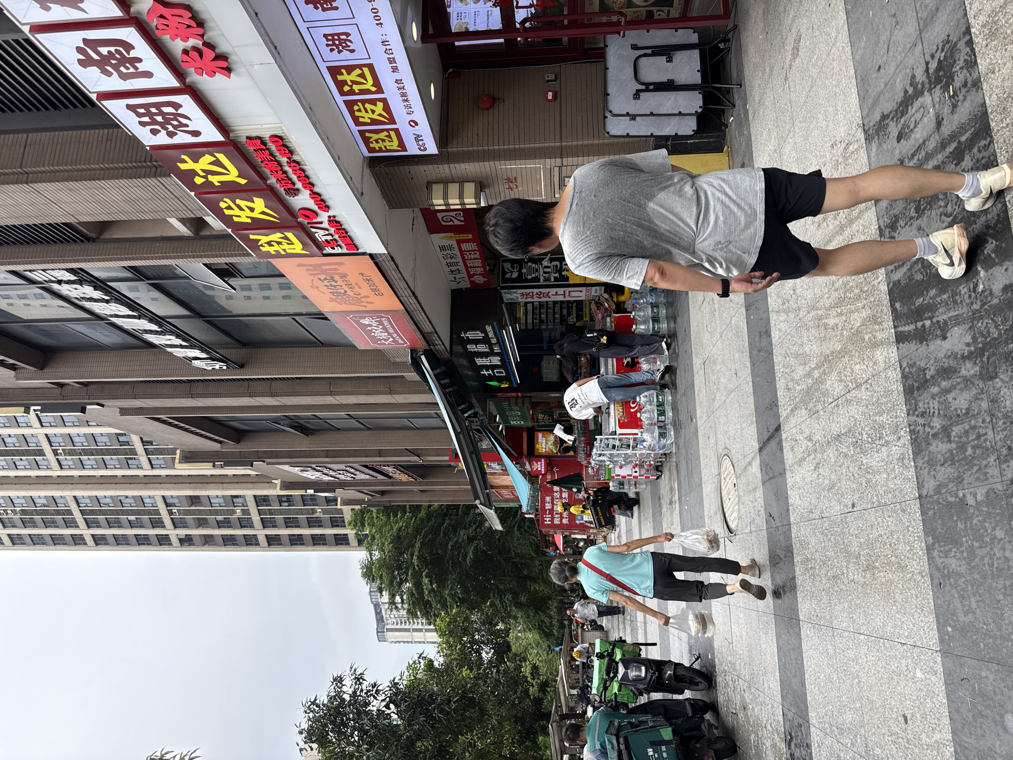 The image shows a street scene in Haizhu District, China, on an overcast day. In the foreground, a man wearing a gray t-shirt and black shorts walks away from the camera on a wet, tiled sidewalk. To the left, a woman wearing a teal shirt and dark pants walks away from the camera carrying two plastic bags filled with food items. She is walking on the same sidewalk as the man. In the background, there are several buildings, including a tall, modern apartment building and a commercial building with storefronts. Several delivery scooters are parked along the sidewalk to the left of the frame. Other people are visible in the background, some of whom appear to be customers or employees of the shops. The storefronts display various signage with Chinese characters, some of which advertise food and beverages. There are also stacks of bottled water and coolers outside one of the shops. The overall environment suggests a bustling urban street with commercial activity.