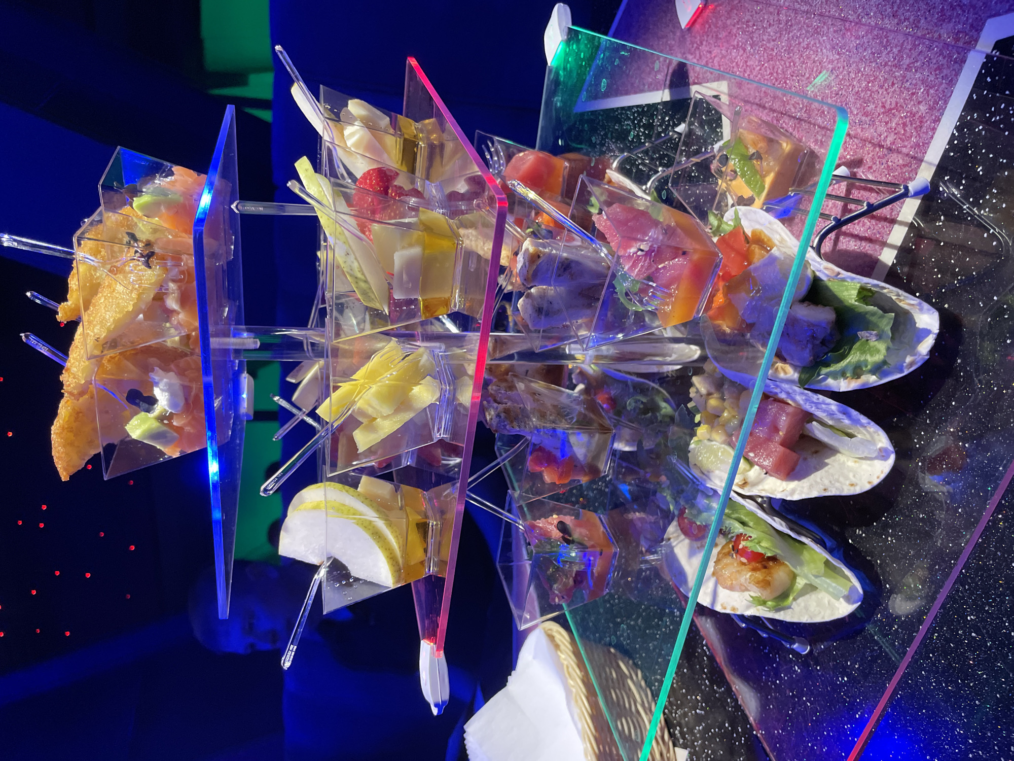 A high-angle, interior shot depicts an elaborate food presentation, likely at a restaurant or event. The presentation involves multiple small, clear containers holding various food items – including sliced cheese, fruit (possibly strawberries), fried food, and other unidentified components – arranged in a multi-tiered structure. The structure is constructed using transparent, colored (blue, red, and green) acrylic or glass panels.
Next to this structure, on a black surface with a speckled or starry pattern, several small tacos are arranged. These tacos contain various ingredients such as lettuce, seafood (possibly shrimp), tuna, and corn. Adjacent to the tacos, a portion of a woven basket and white napkin is visible.
The background is blurred, with dark blue and pink hues, suggesting an artificially lit environment. Based on the image, there is no way to ascertain its location as Saint Petersburg, Russia.