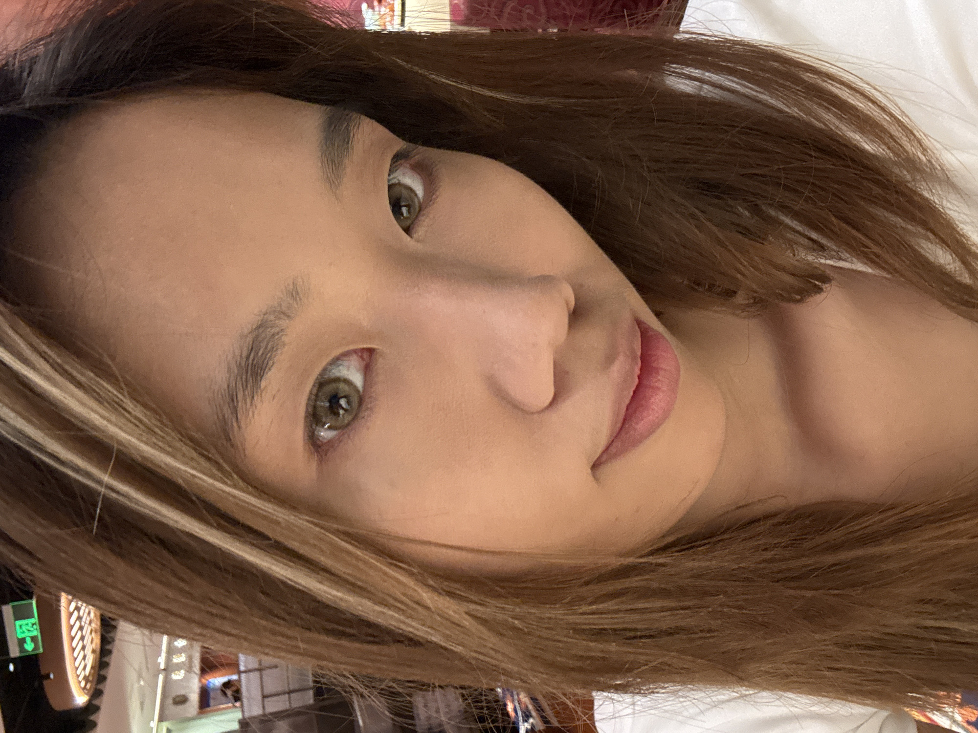 A close-up selfie of a young East Asian woman with light brown hair, partially covering her face. Her eyes are a light green color. She is wearing a white top. The background is blurred, but appears to be an indoor commercial setting with some signage that may indicate a location in Jiulongpo District, China. There are overhead lights and other people visible in the distance.