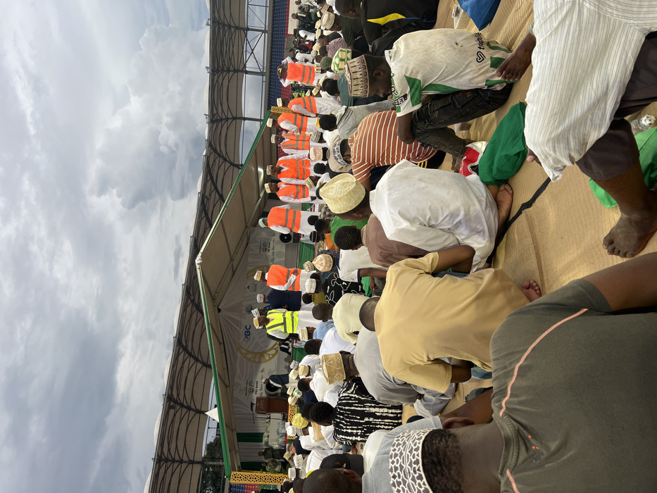 The image captures a wide-angle view of a large outdoor gathering of predominantly men at Amaan Stadium in Zanzibar City, Tanzania. The attendees are seated cross-legged on light brown woven mats, facing an elevated stage in the background. Many individuals wear traditional kufi caps, varying in color and pattern (white, beige, brown, black).
The stage is covered by a large white tent structure with green trim, featuring the "PBZ BANK" logo, a crescent moon and star motif, and the text "The People's Bank, For the People." On the stage, a dark wooden podium with a microphone is visible. Several men are present on or immediately in front of the stage; some wear white thobes and kufis, while others wear high-visibility safety vests (orange or yellow) over their clothing. One individual in a suit is visible behind the podium.
Beyond the stage structure, the curved, open-air upper tiers of stadium seating, colored blue, are discernible. The sky above is heavily overcast with dark grey clouds. The scene portrays a formal, large-scale event or ceremony.