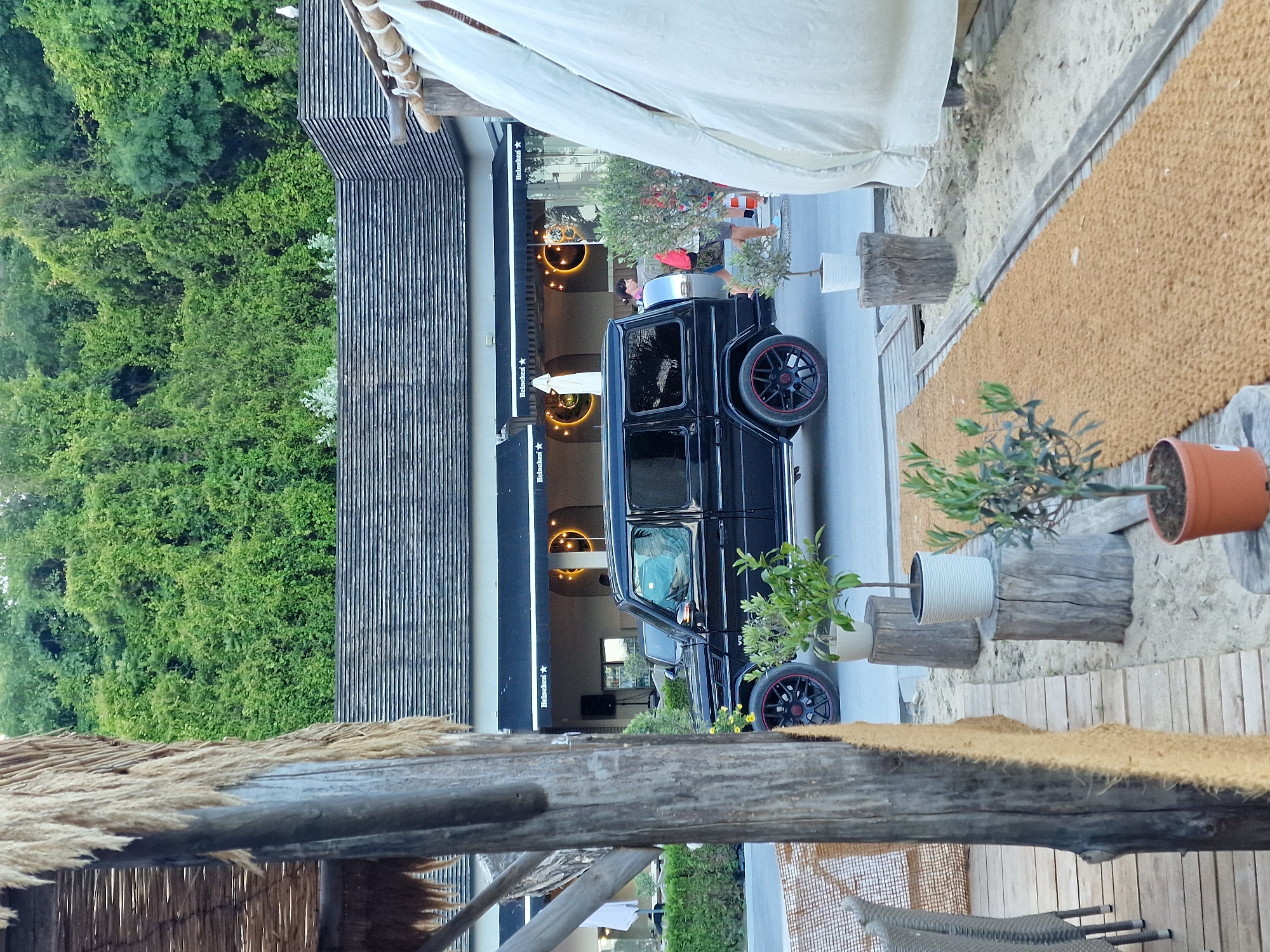 In an outdoor setting with a beach-like environment, a black Mercedes-Benz G-Class SUV is parked on a paved road. The vehicle has black rims with red accents. In the background, a building with a dark, sloped roof is visible, featuring awnings with "Heineken" logos. Dense green trees and vegetation form a backdrop behind the building. To the right of the SUV, people are walking; one individual is carrying a red bag. Potted plants are placed on wooden stumps on the sandy ground near the road. In the foreground, a thatched-roof structure with a wooden pillar partially obscures the view of a table and chairs. White fabric is draped on a structure to the far right. The scene appears to be a coastal establishment, possibly a restaurant or bar.