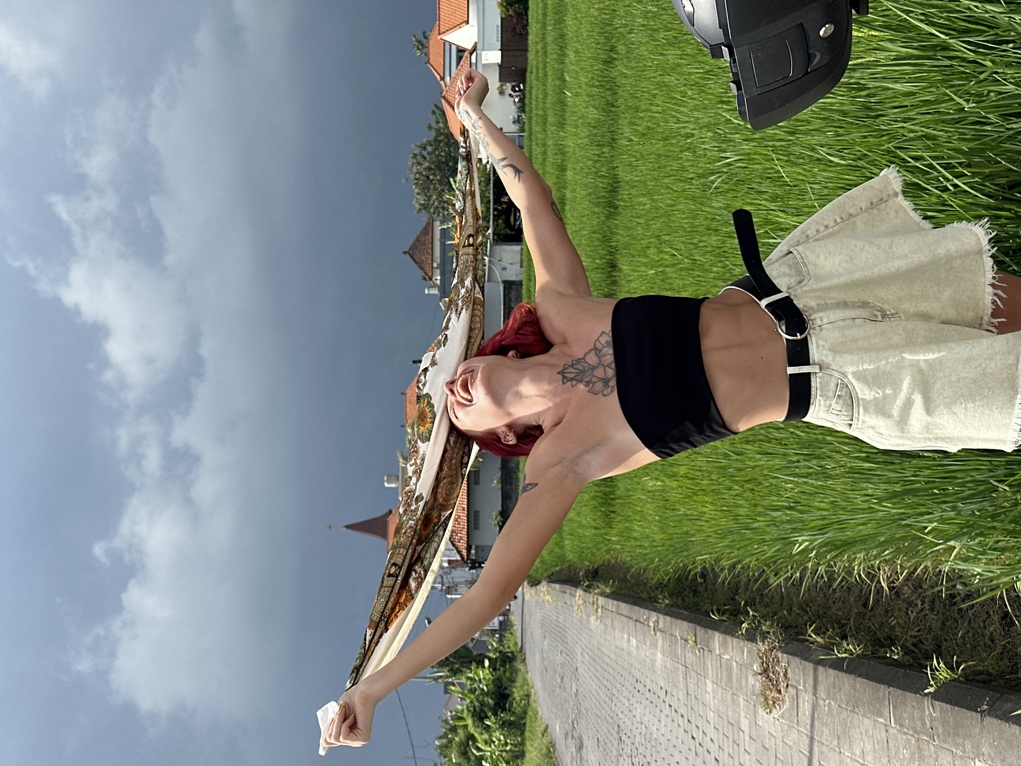 A young woman with red hair, dressed in a black crop top and light-colored denim shorts with a black belt, is captured in a medium shot outdoors. She is holding a patterned scarf above her head with both hands extended. The scarf features a cream background with green, brown, and orange details.
The background shows a grassy area, a paved sidewalk, and several buildings with orange tile roofs typical of architecture found in the city of North Kuta, Indonesia. A dark sky with stormy clouds hangs overhead, contrasting with the bright green of the grass and the warmer colors of the buildings. A partial view of a motorcycle helmet is visible in the upper right corner of the frame. The photograph is taken at a slight angle, rotated clockwise.