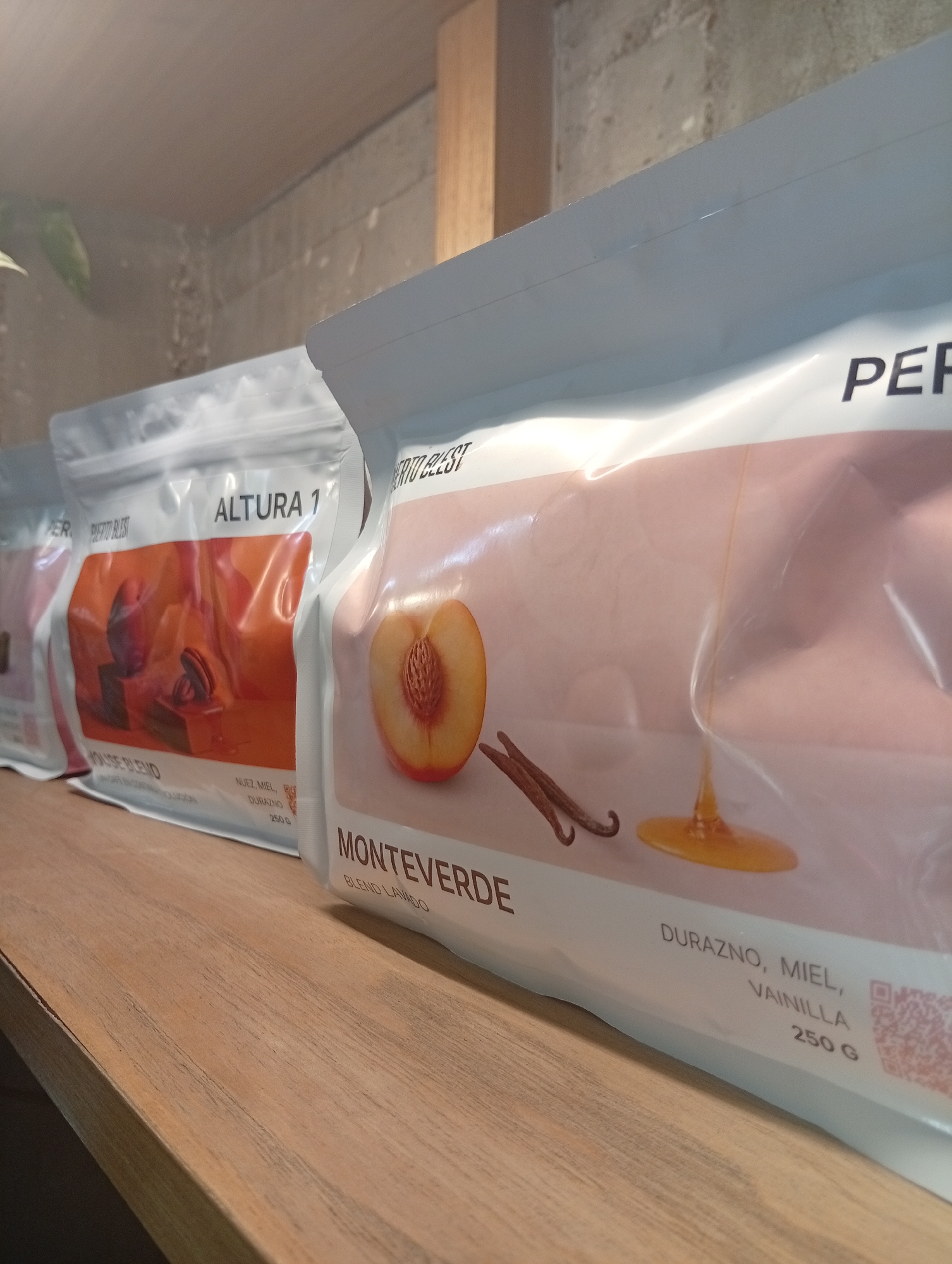 The image shows a close-up of three retail coffee packages from the "PER PIERTO BLEST" brand, arranged in a row on a wooden shelf. The package closest to the viewer is labeled "MONTEVERDE" and features a peach, vanilla beans, and a honey droplet on its design. The next package is labeled "ALTURA 1" and has an illustration of plums, a bean grinder, and coffee beans. The shelf appears to be located inside a store that specializes in coffee. The packages list "DURAZNO, MIEL, VAINILLA" (peach, honey, vanilla) as flavor notes, and the product weighs 250 grams. The store may be located in Buenos Aires, Argentina, given the possible Spanish on the product packaging.
